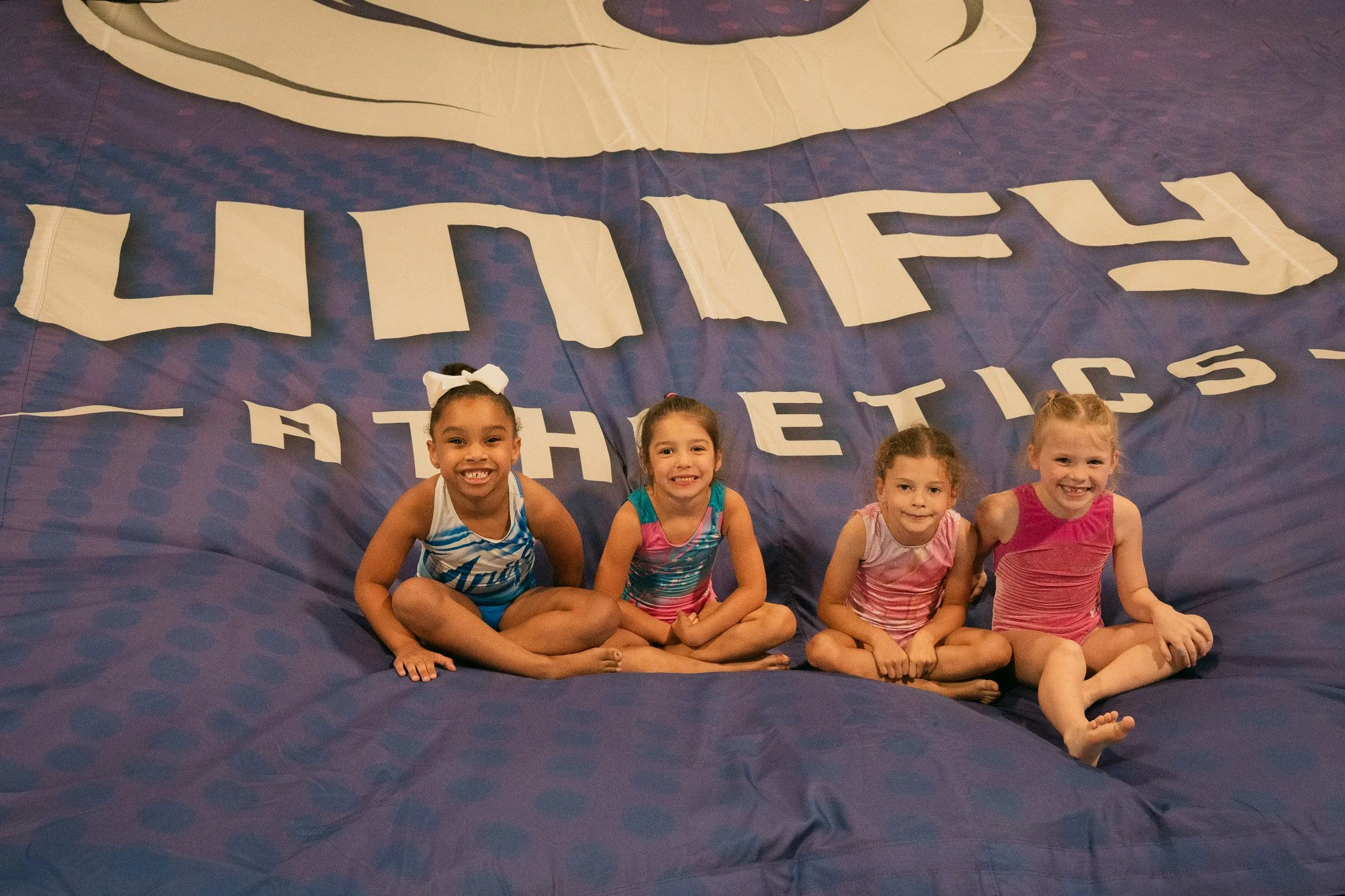 Four young girls sitting on a large blue gymnastics mat with the words 'UHS ATHLETICS' printed on it, smiling at the camera.