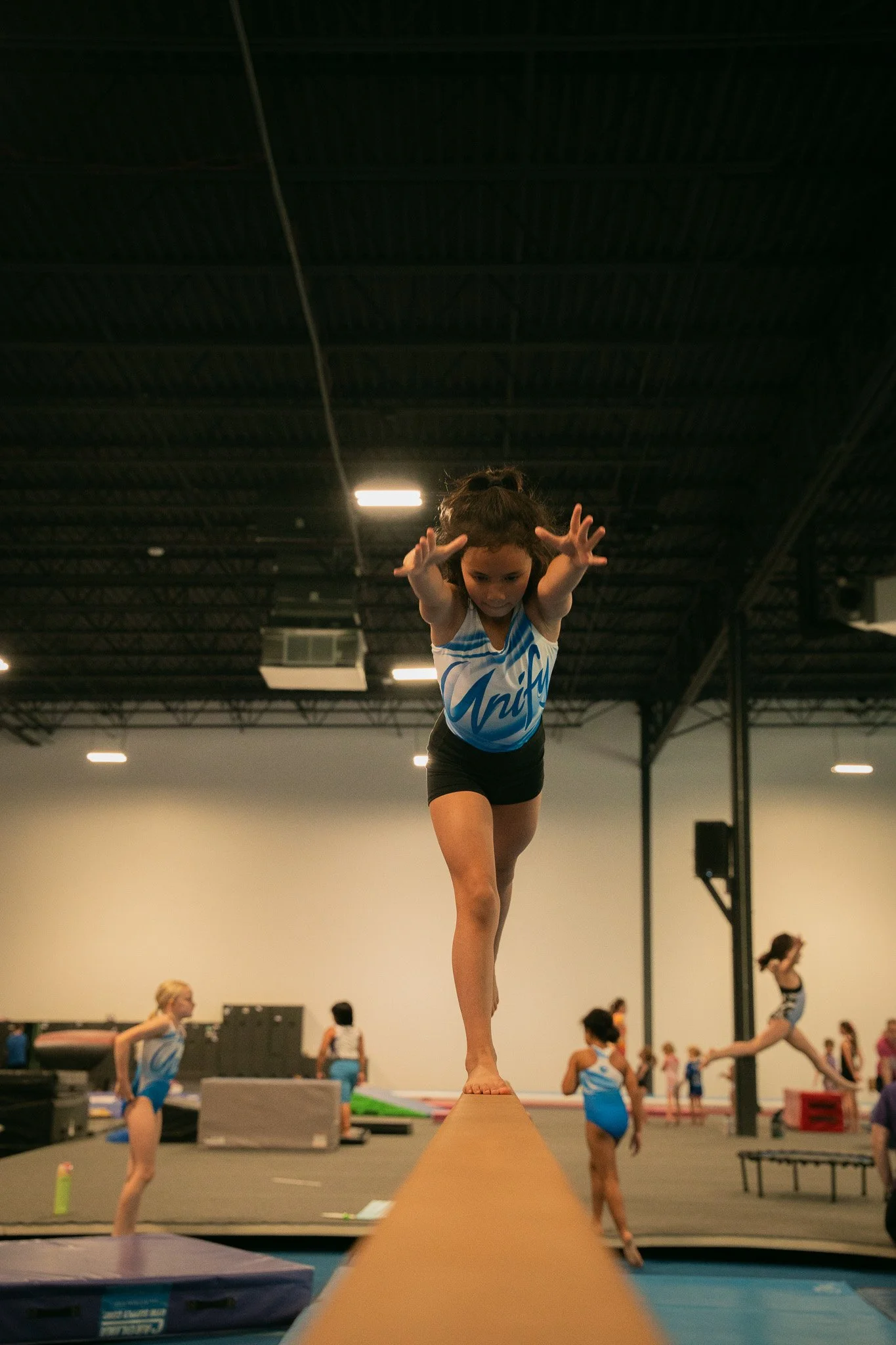 Young girl in gymnastics uniform balancing on a beam in a gymnasium with other children practicing in the background.
