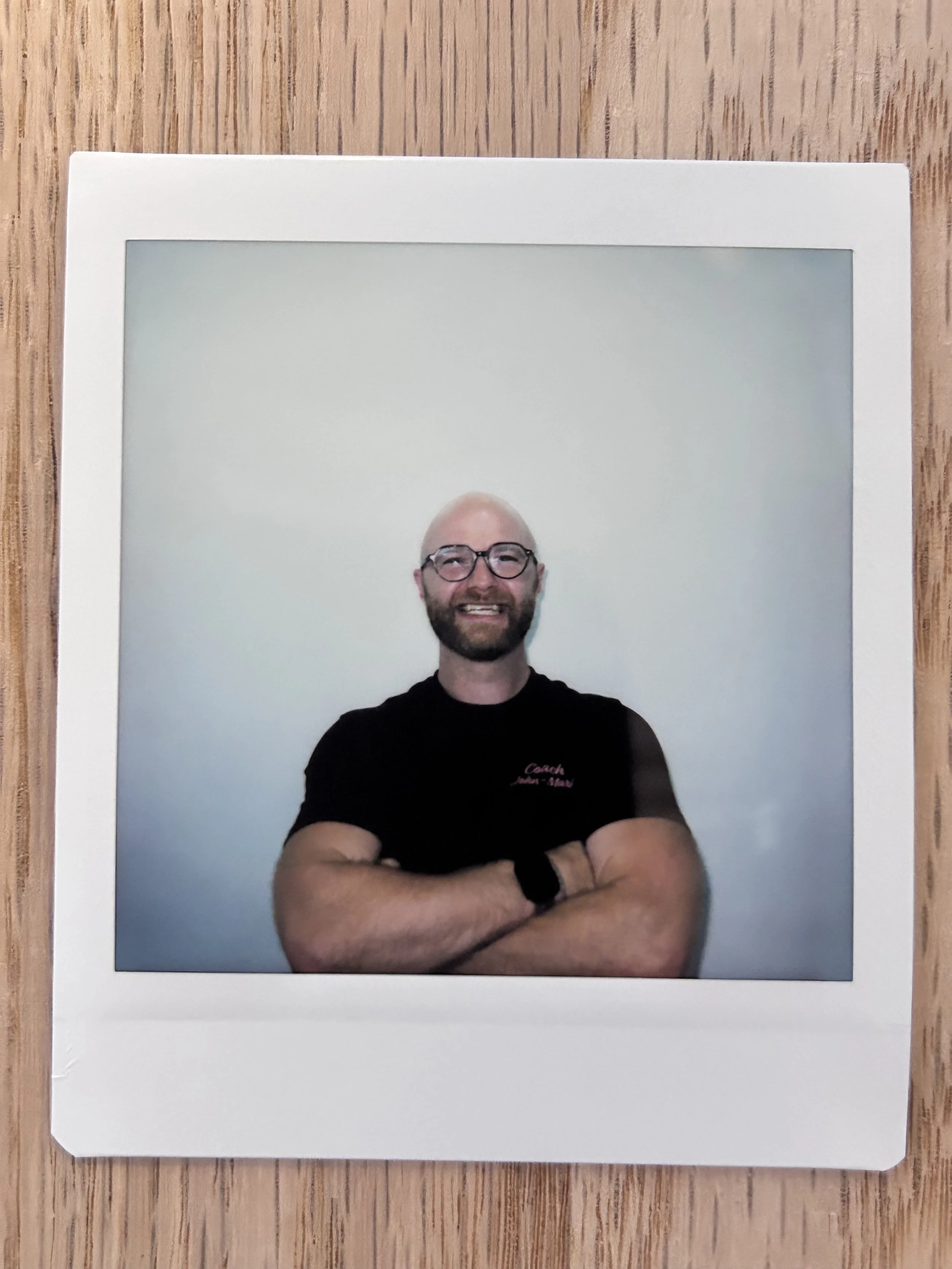 A smiling man with glasses, a beard, and a black t-shirt with red text, standing with arms crossed against a plain light-colored wall.