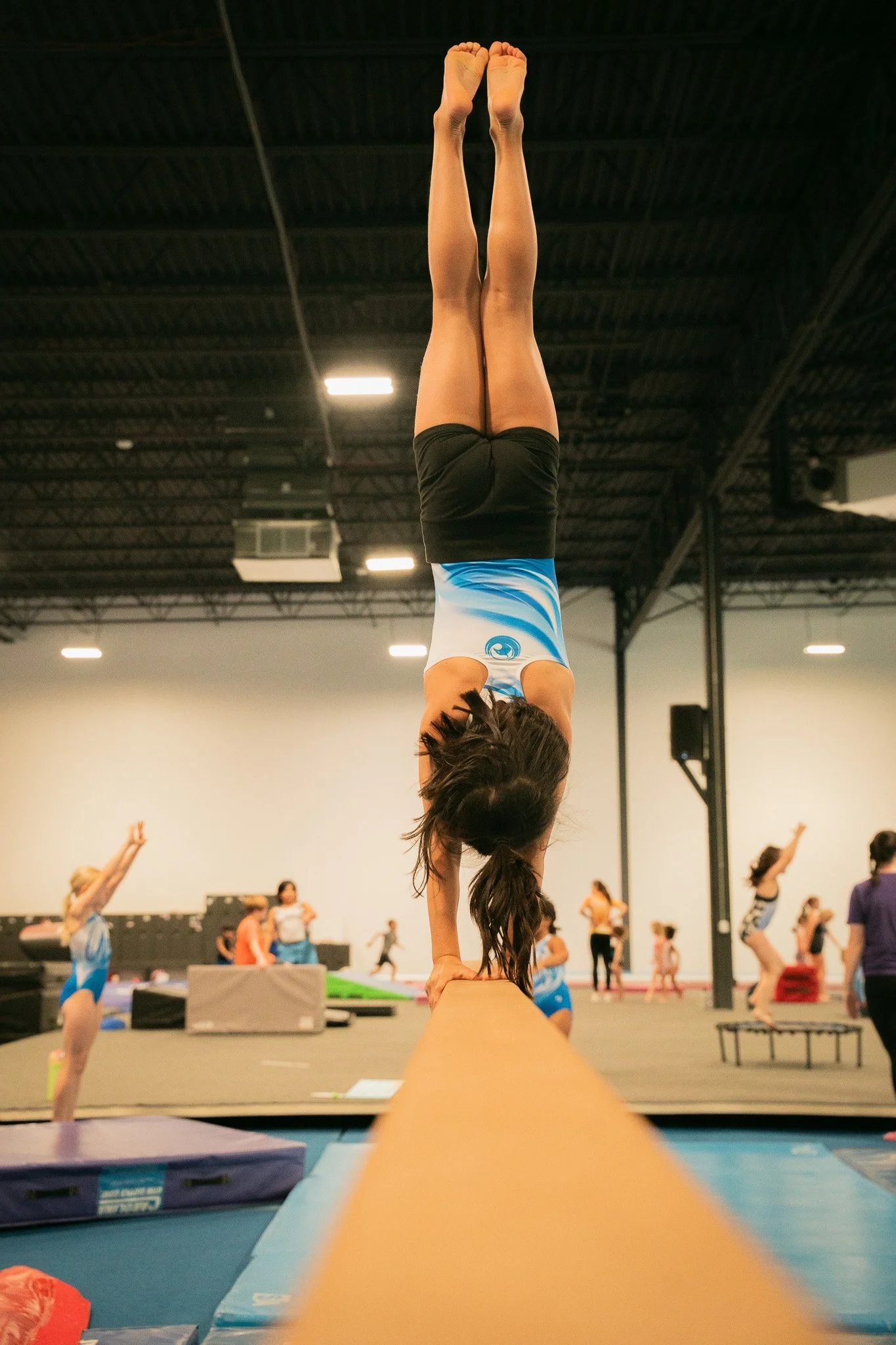 A young gymnast practicing a handstand on a balance beam in a gymnasium, with other gymnasts in the background.