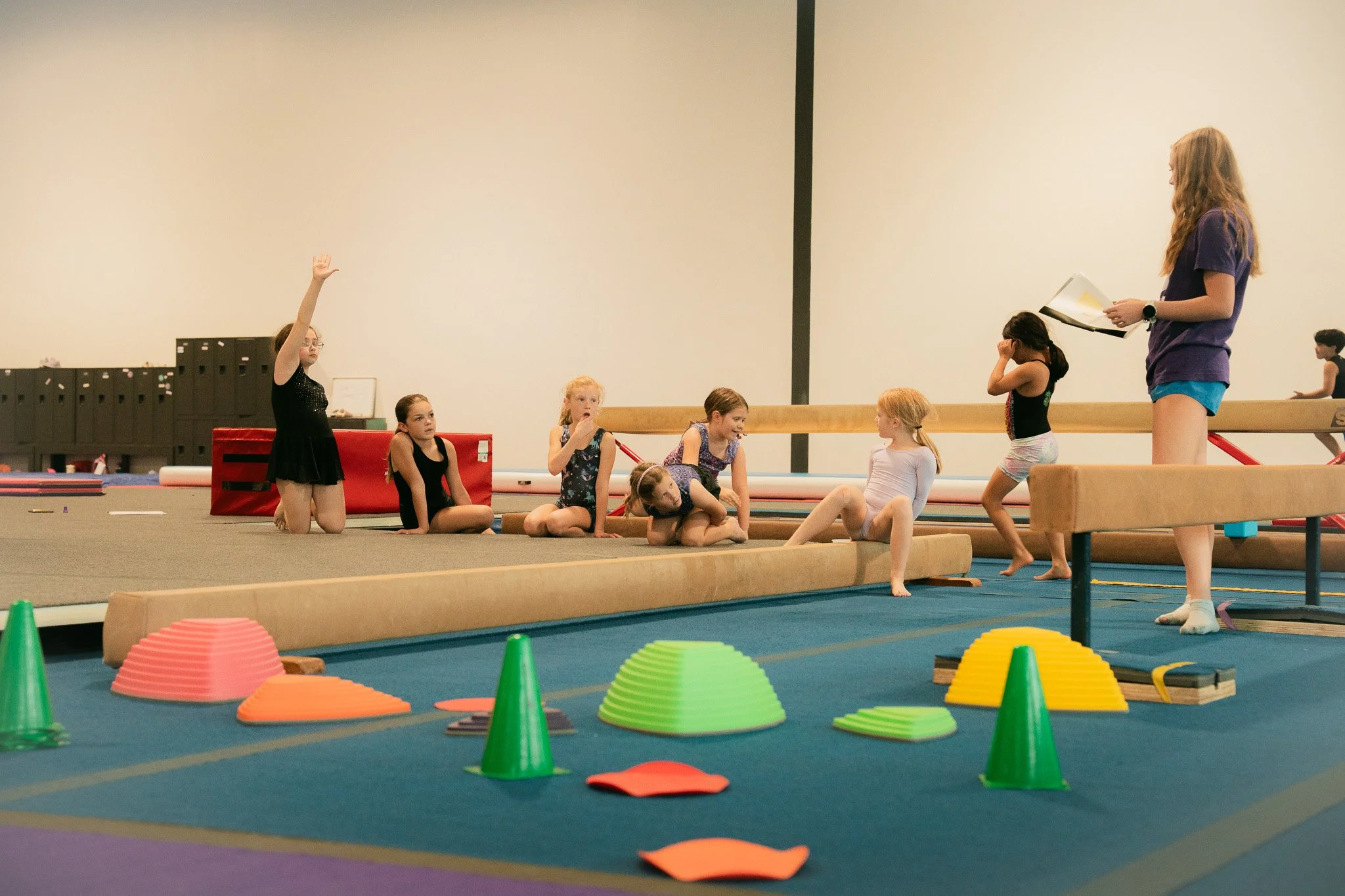 Kids at a gymnastics class listening to instructor, some sitting, one raising hand, gymnastic equipment and cones on floor.