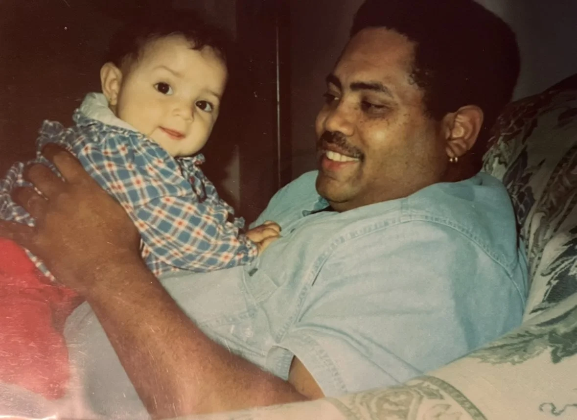 A man with short dark hair and a goatee lying on a bed, smiling and holding a young child. The child has curly dark hair, light skin, and is wearing a plaid shirt. The man has medium skin tone, and the background shows a pillow and dark wall.