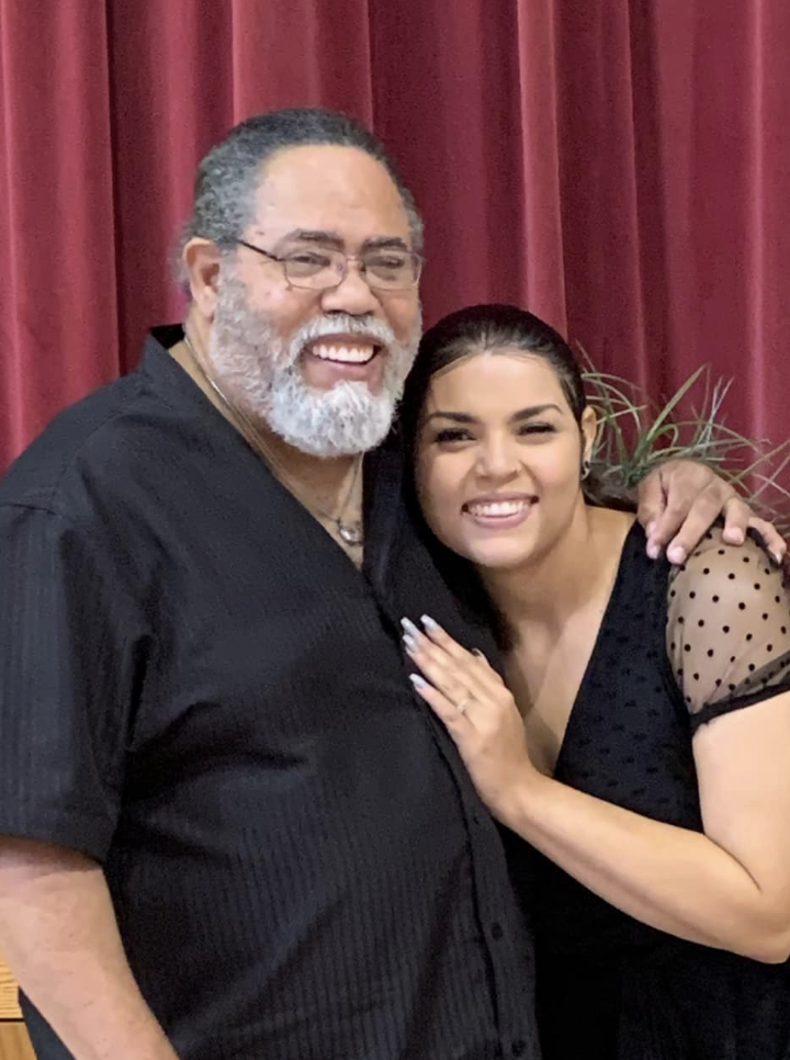 A smiling older man with glasses and a white beard standing next to a smiling young woman, both hugging. They are indoors in front of a red curtain and a potted plant.
