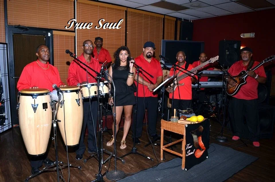 A band called 'True Soul' performing indoors with seven musicians, including percussion, congas, vocals, guitar, and keyboard, all wearing red shirts, with a woman singer in the center.