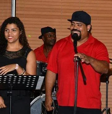 A man with a beard and mustache wearing a black cap and red shirt, holding a microphone, standing next to a woman with curly hair wearing a black dress, and another man in a red shirt in the background playing drums, indoors with wooden paneling.