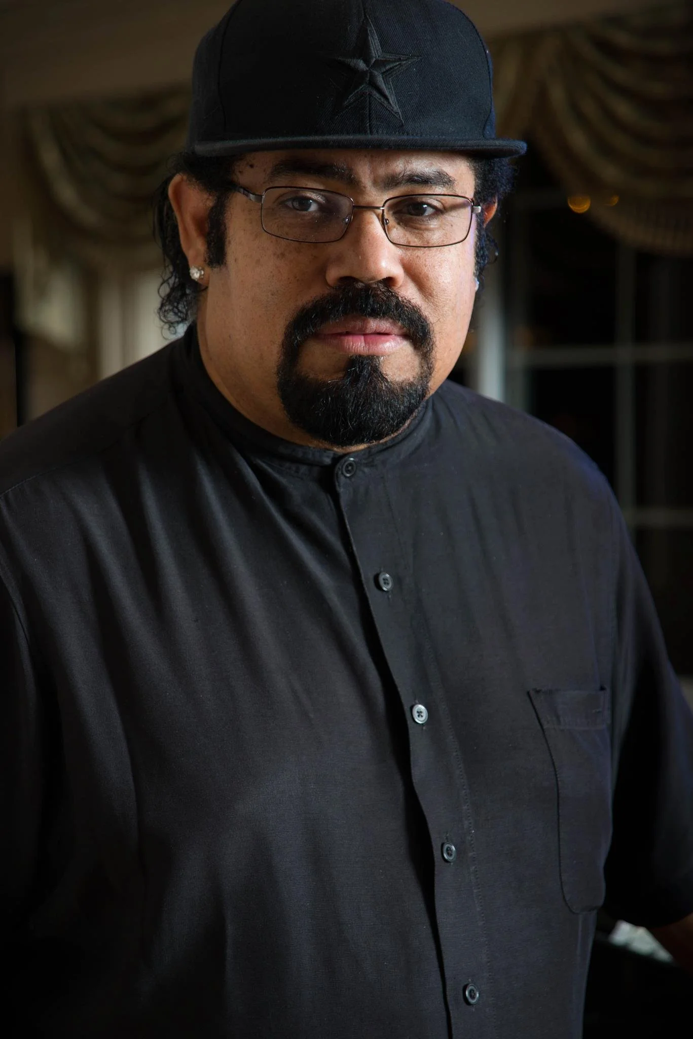 A man with a mustache and goatee wearing glasses, a black button-up shirt, and a black baseball cap with a star emblem. He has earrings and is in an indoor setting with draped curtains in the background.