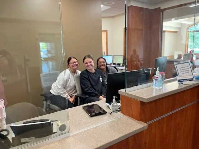 Three women sitting at a reception desk, smiling, in an office environment with computers and a glass partition.