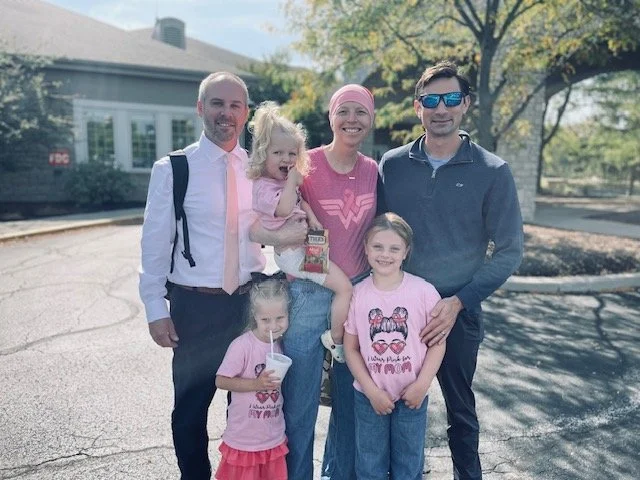 A family of five, two adults and three children, posing outdoors on a sunny day. The adults are men and women, and the children are two girls and a young girl. They are standing on a paved area with a building and trees in the background.