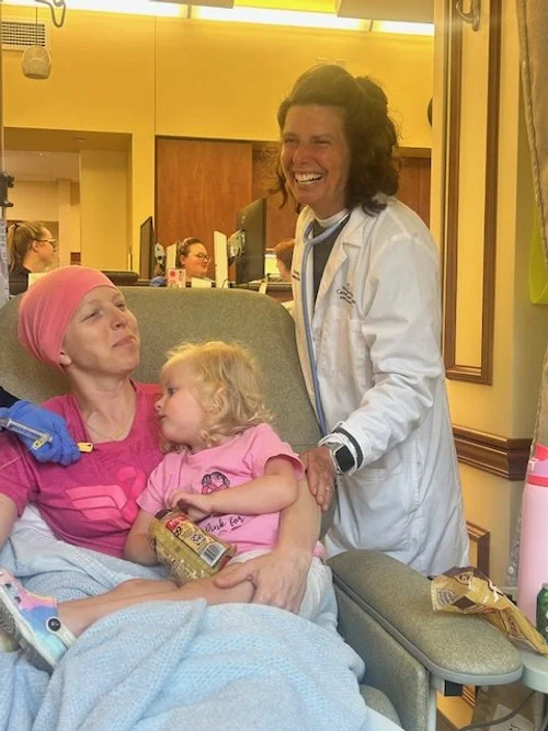 A woman with a brain tumor, wearing a pink headscarf and hospital gown, sitting on a hospital bed with a young girl, who is also in a hospital gown, holding a snack. A medical professional in a white coat is standing nearby, smiling and talking with 