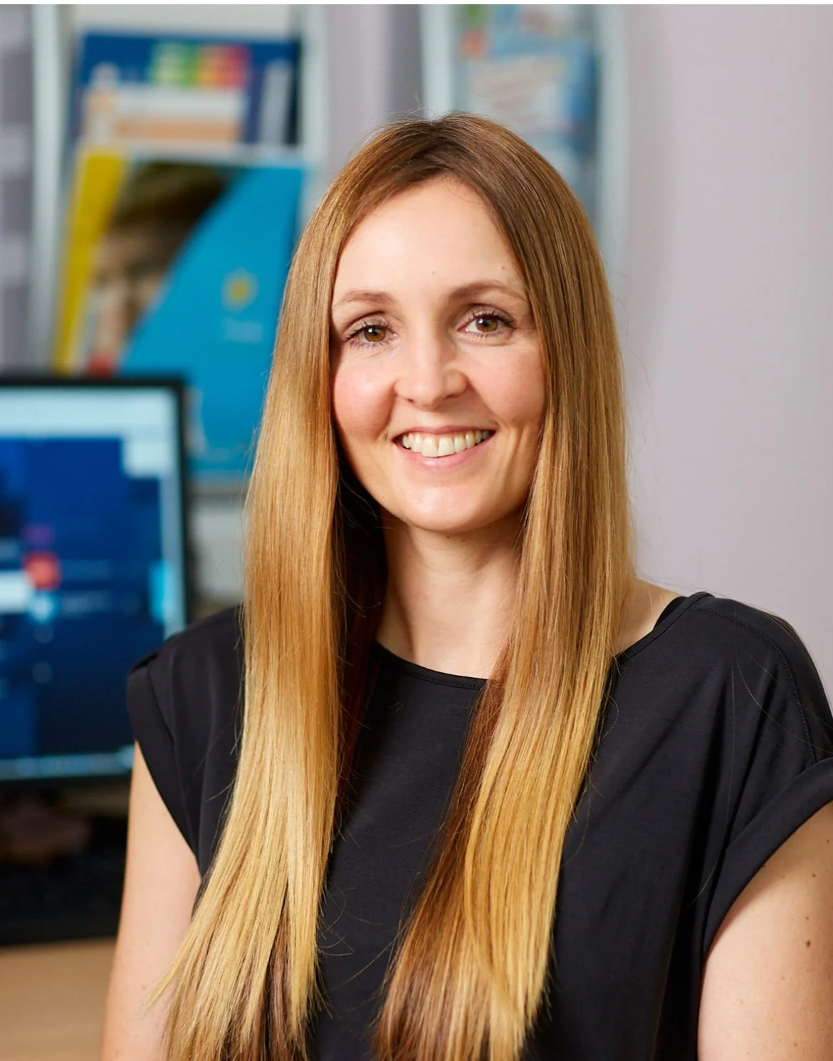 A woman with long red hair smiling in an office environment with computers and books in the background.