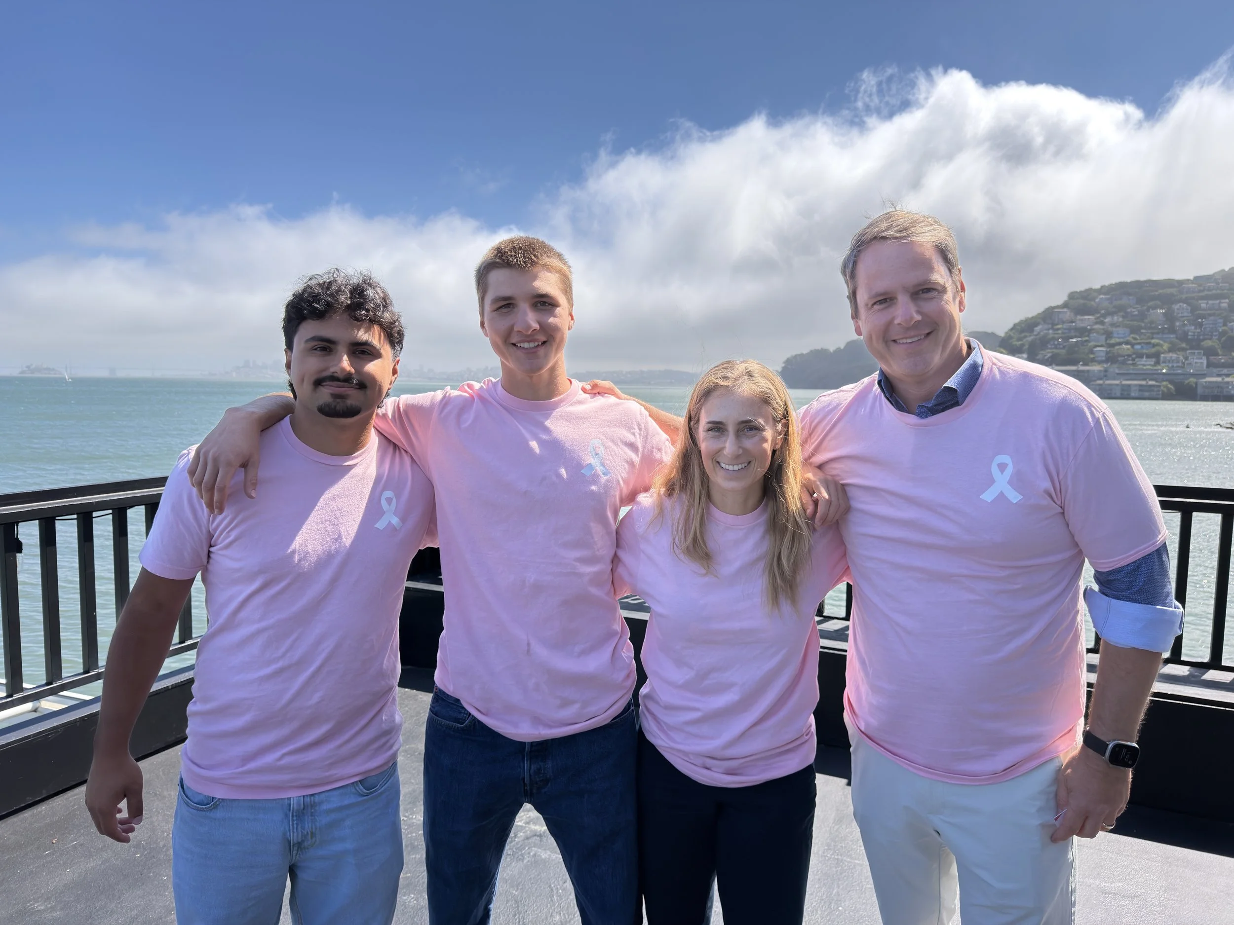 A group of four people, two men and two women, standing together outdoors on a railing with water and a hilly residential area in the background. They are all smiling and wearing pink shirts with a light blue awareness ribbon. The sky is partly cloudy.