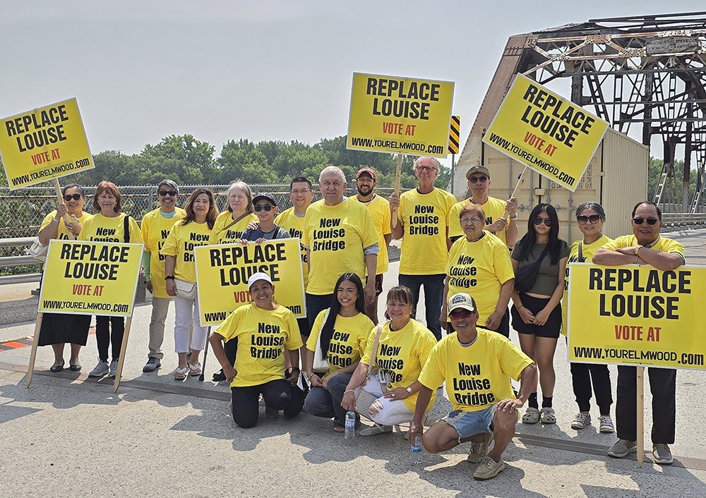 Group of people holding signs that say "Replace Louise" and "Vote at www.yourelmwood.com," wearing yellow shirts with "New Louise Bridge" printed on them, standing outdoors near a bridge.