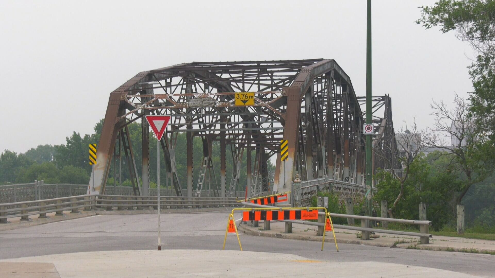 Old rusted metal bridge with signs indicating a height limit of 3.76 meters, partially blocked off with caution tape and barricades, surrounded by trees and cloudy sky.