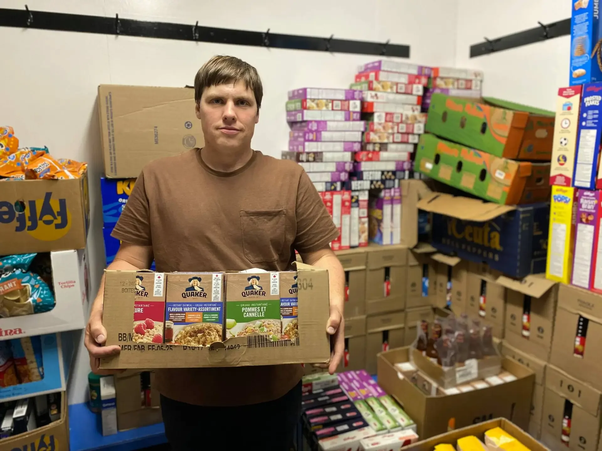 A man wearing a brown t-shirt holding a box of instant food varieties in a storage room filled with boxes of snacks and cereal.