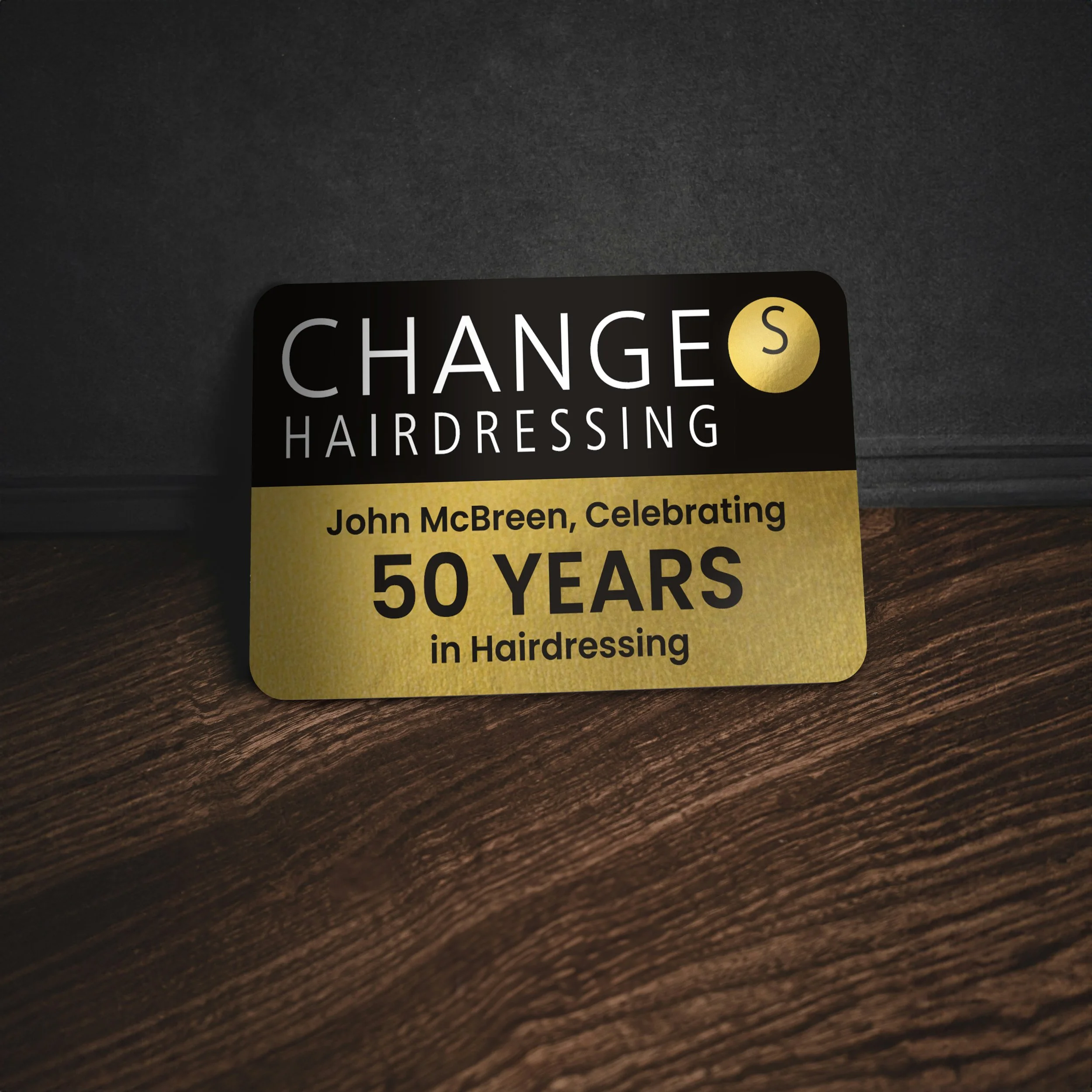 A black and gold sticker celebrating John McBreen's 50 years in hairdressing, placed on a wooden surface.