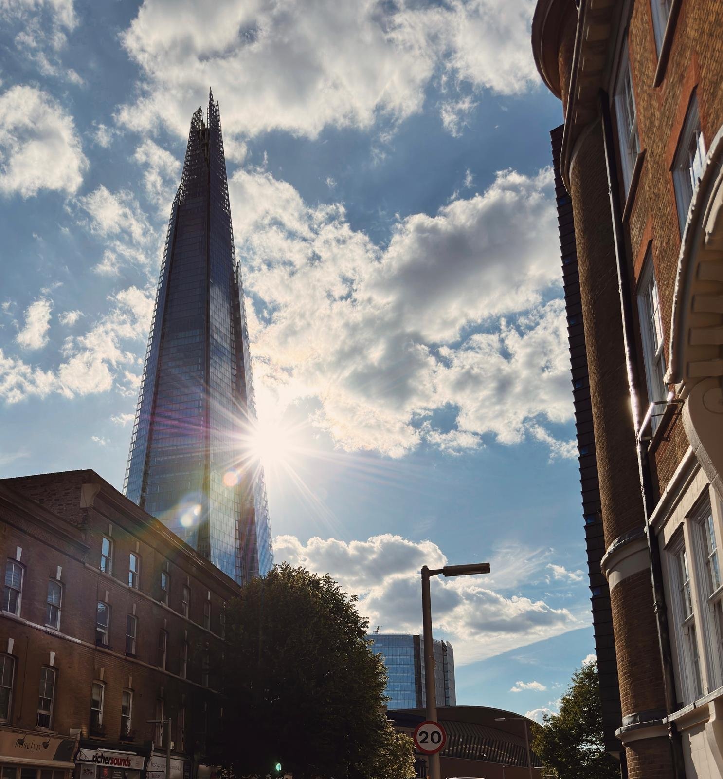 A tall skyscraper (The Shard) with reflective glass windows, partly obscured by clouds and sun rays, in London, surrounded by older buildings and trees.
