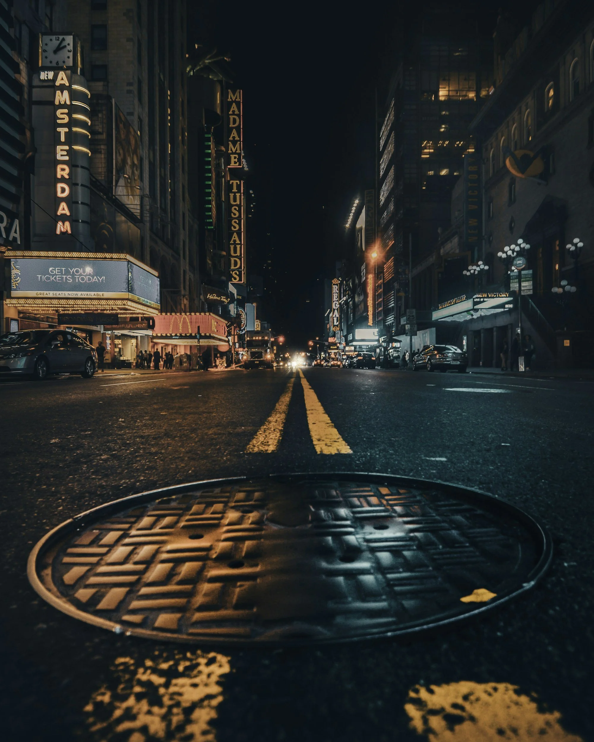 Nighttime city street in Times Square, New York City, with illuminated signs for 'Madame Tussaud's' and 'New Amsterdam Theatre,' cars parked along the sides, and a manhole cover in the foreground.