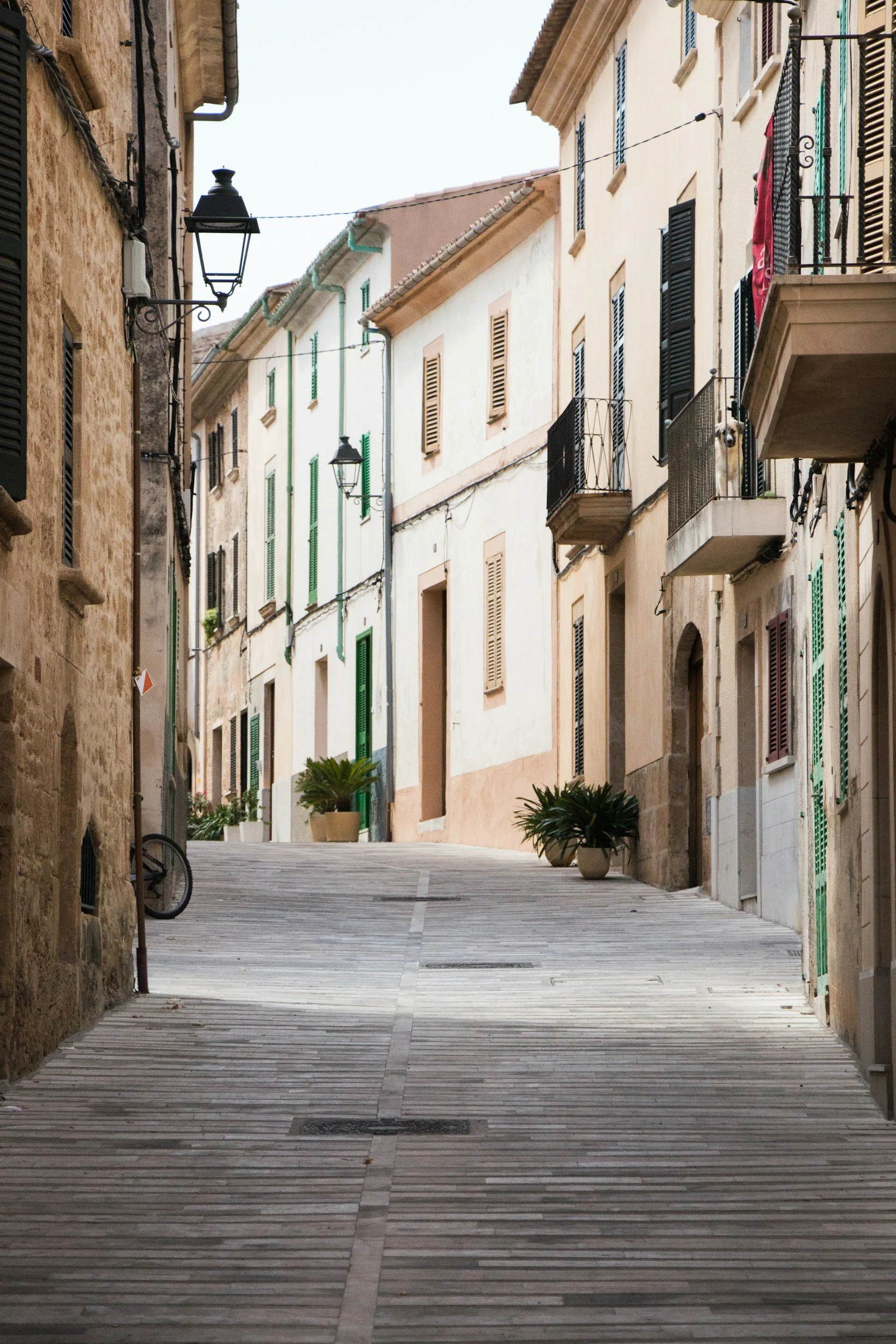 A narrow, sloped street in a European town with old buildings, each with balconies and shutters, and potted plants, lit by vintage-style street lamps.