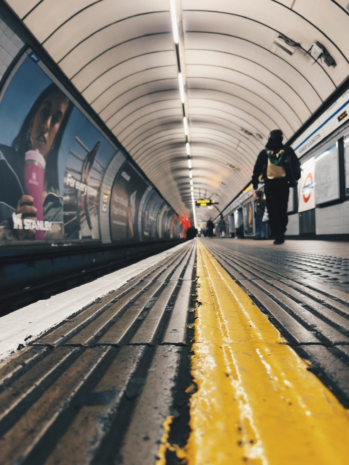 Low-angle view of a subway station platform with a yellow safety line, a person walking with a backpack, and advertisements on the wall.