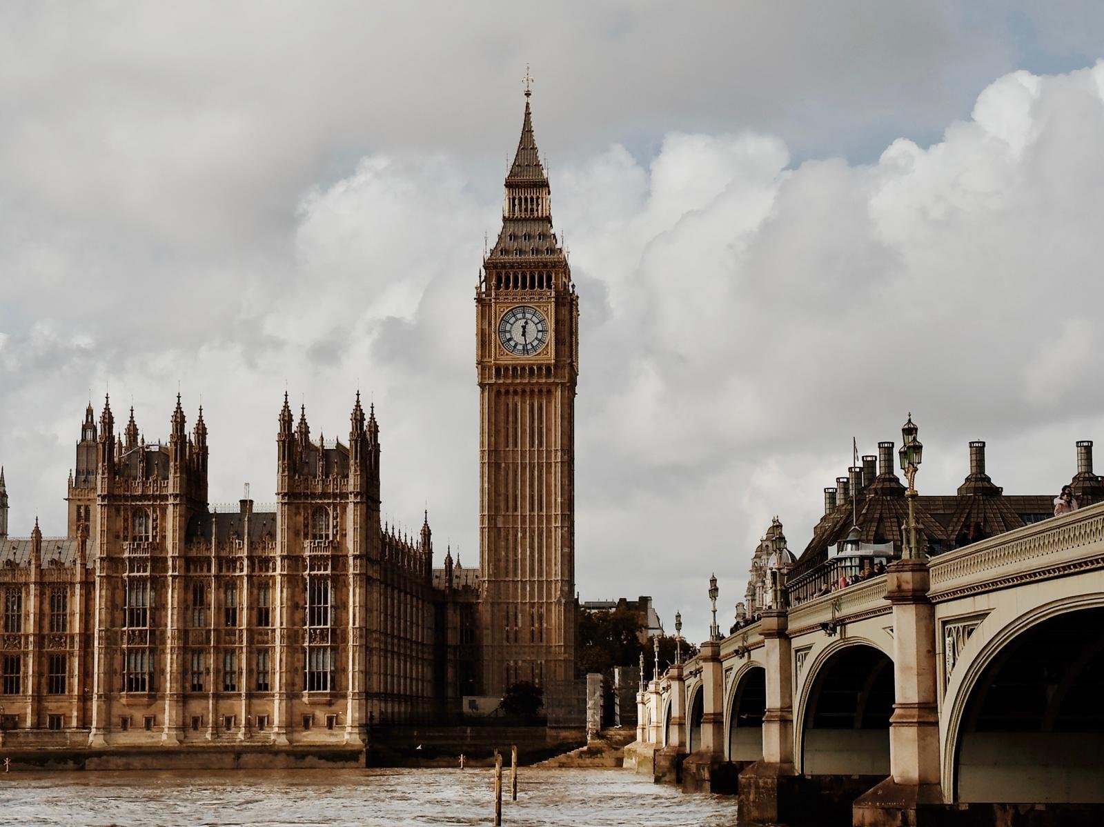 View of the Big Ben clock tower and the Palace of Westminster in London, England, with a bridge over the River Thames in the foreground and cloudy sky overhead.