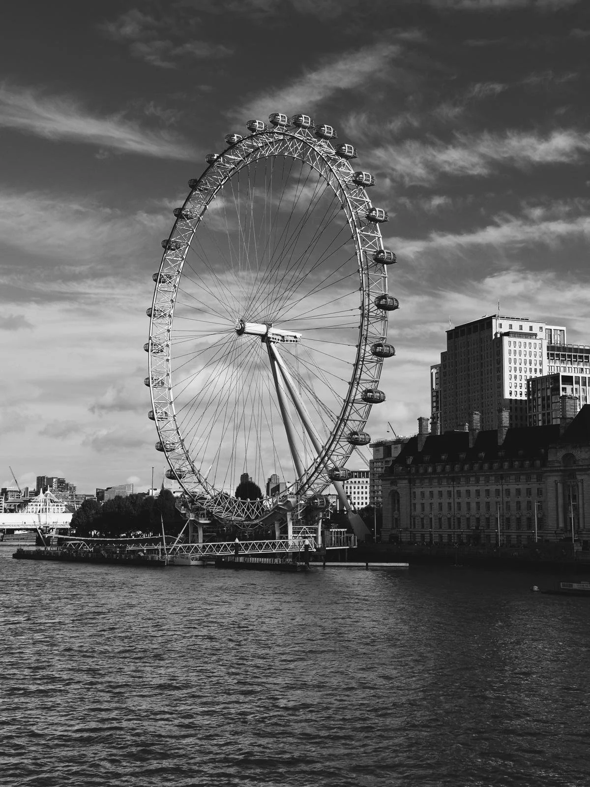 A black and white photo of a large Ferris wheel by the water with city buildings in the background.