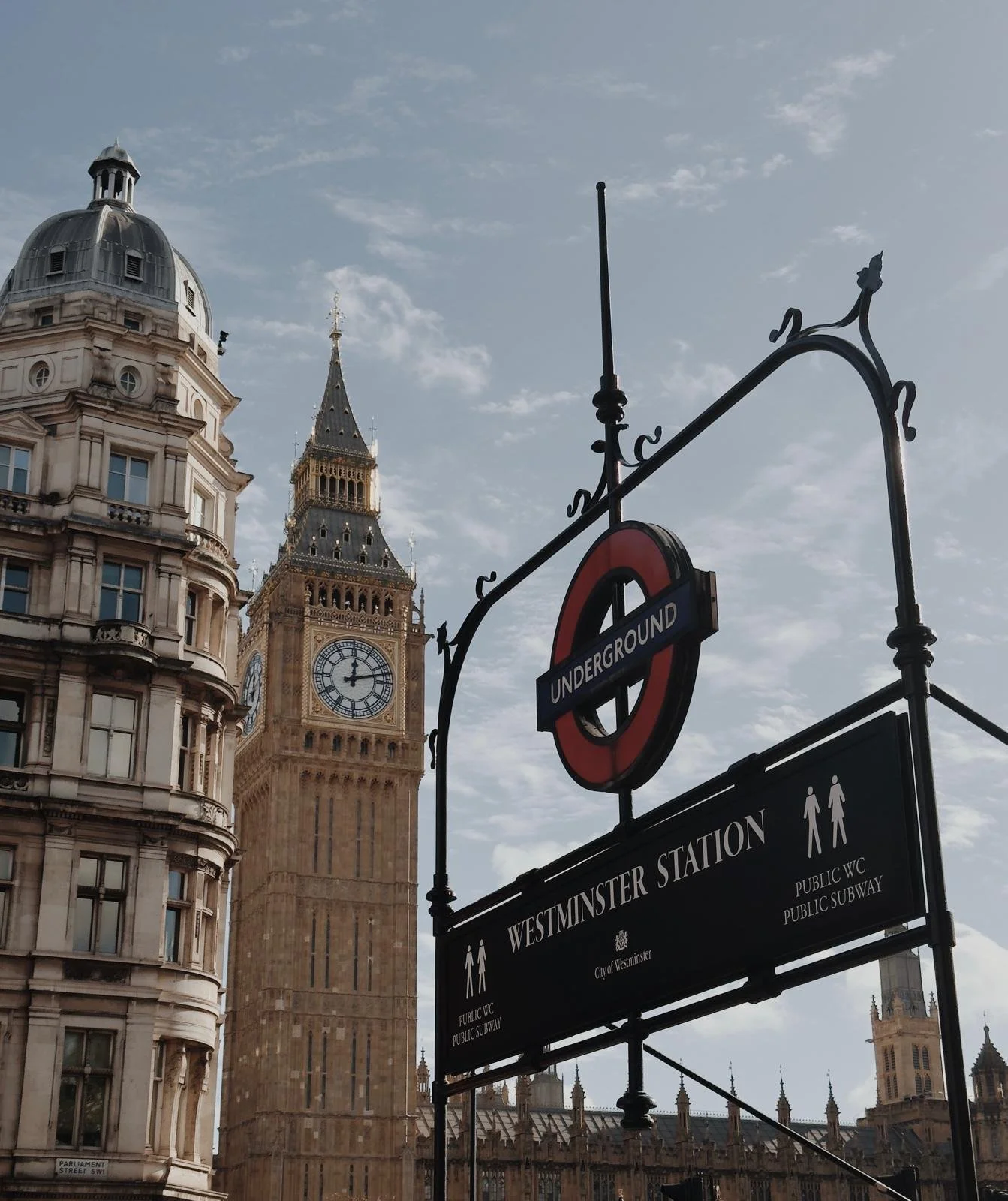 Big Ben clock tower beside Westminster station sign at a London street with historic buildings and partly cloudy sky in the background.