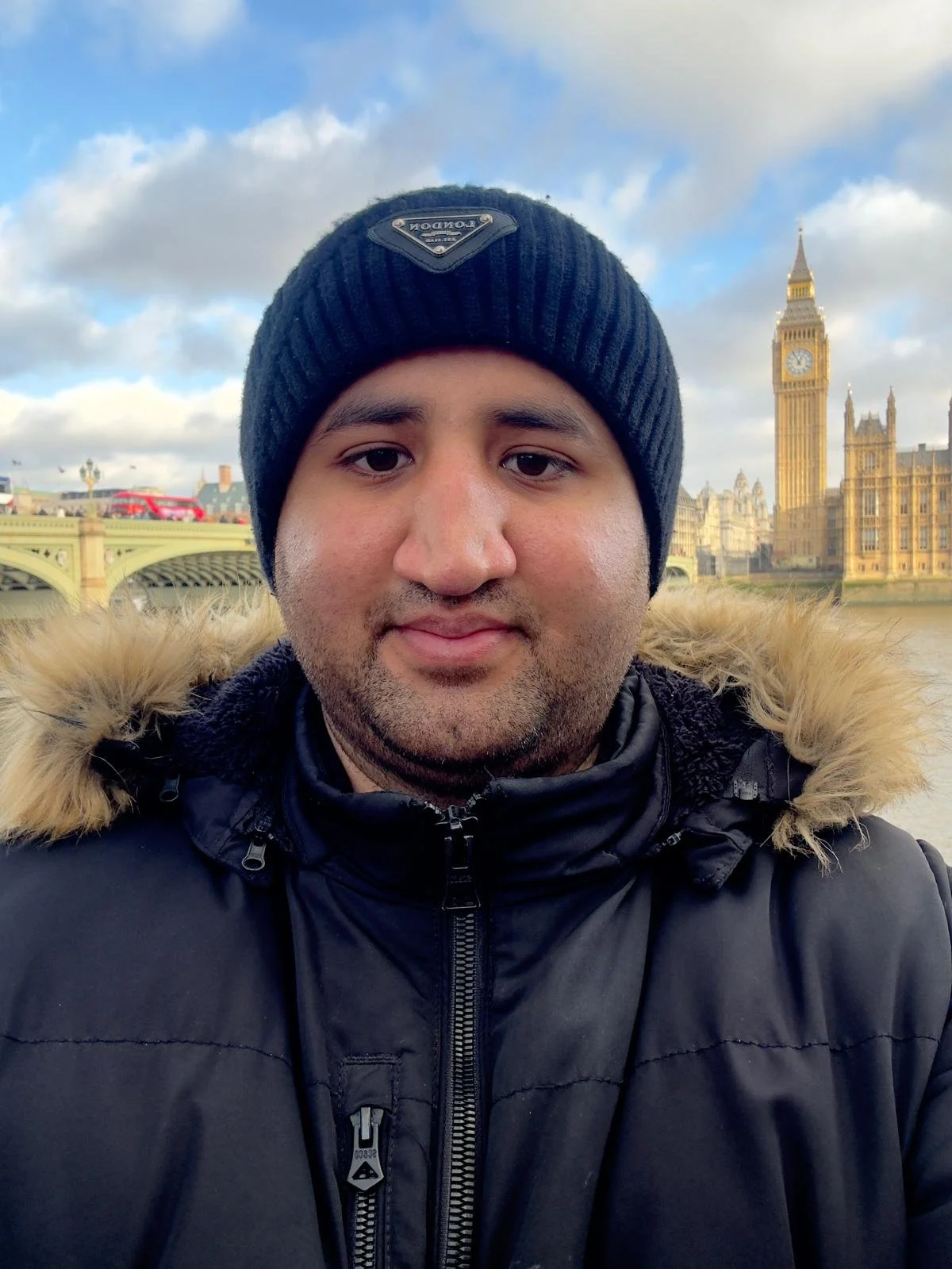 A man wearing a black beanie and a black winter jacket standing in front of the River Thames with the Westminster Palace and Big Ben in London in the background.