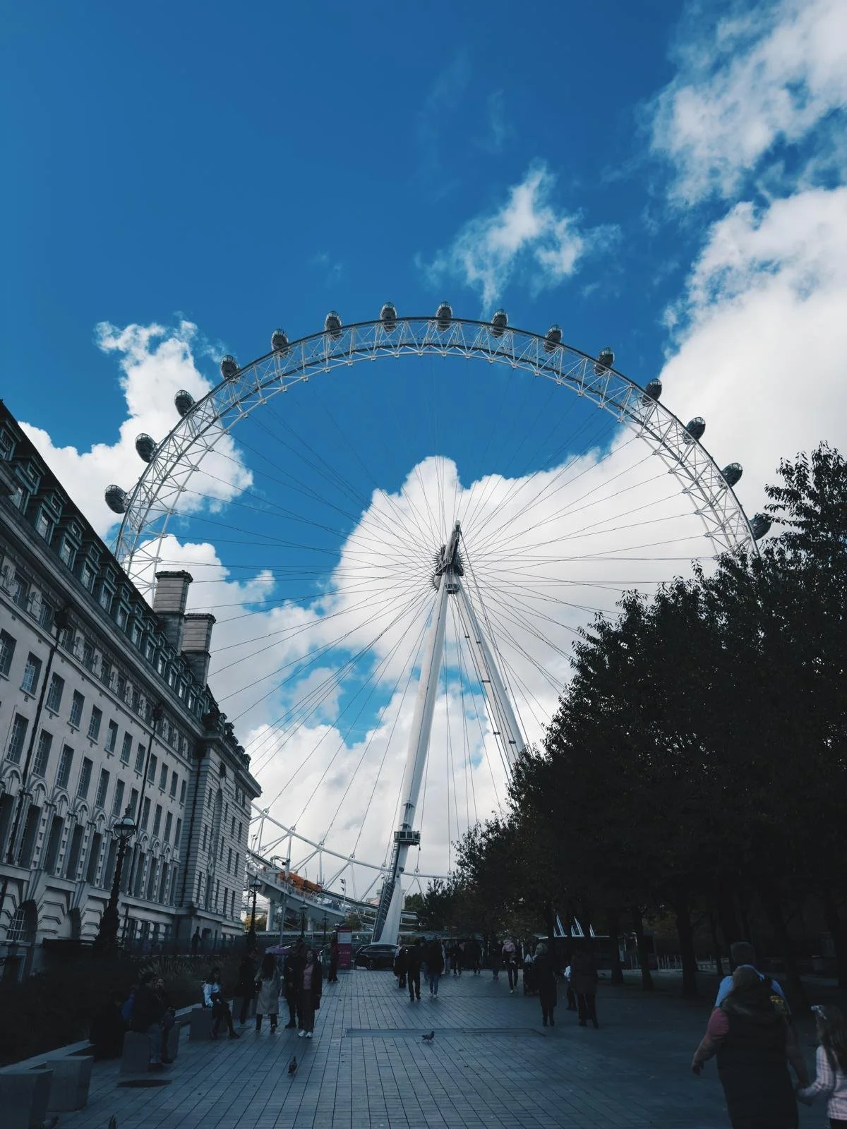 London Eye Ferris wheel against a partly cloudy sky, located near historic buildings and a crowd of people walking on a paved walkway.