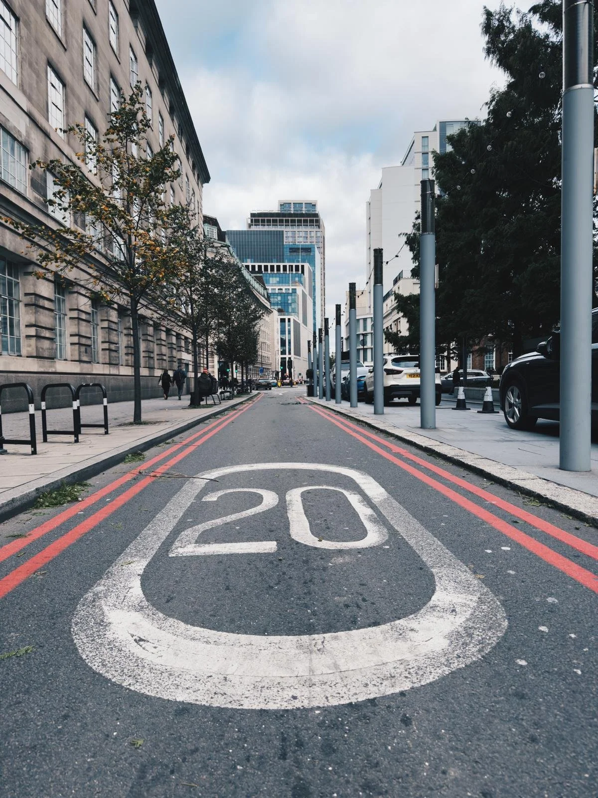 City street with 20 mph speed limit marking on the road, parked cars, tall buildings, trees, and pedestrians.