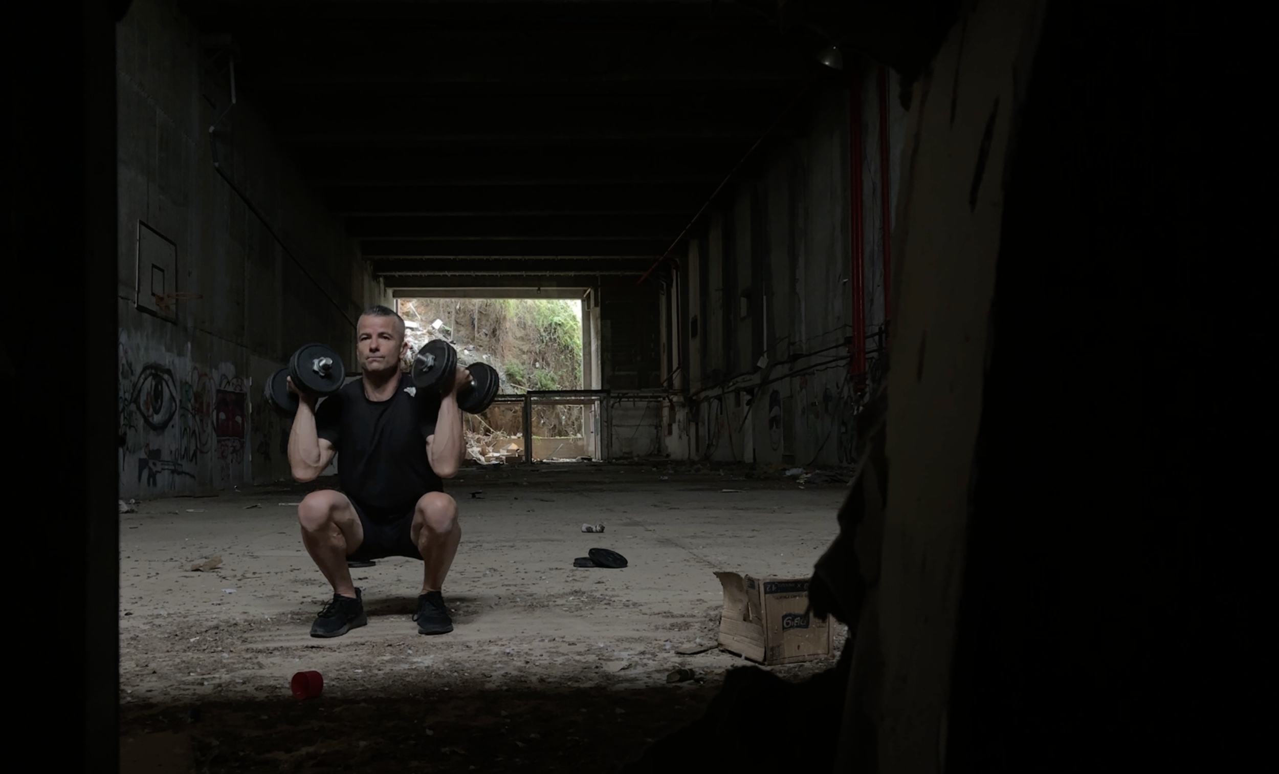 A man is exercising with dumbbells in an abandoned, graffiti-covered tunnel with debris on the ground.