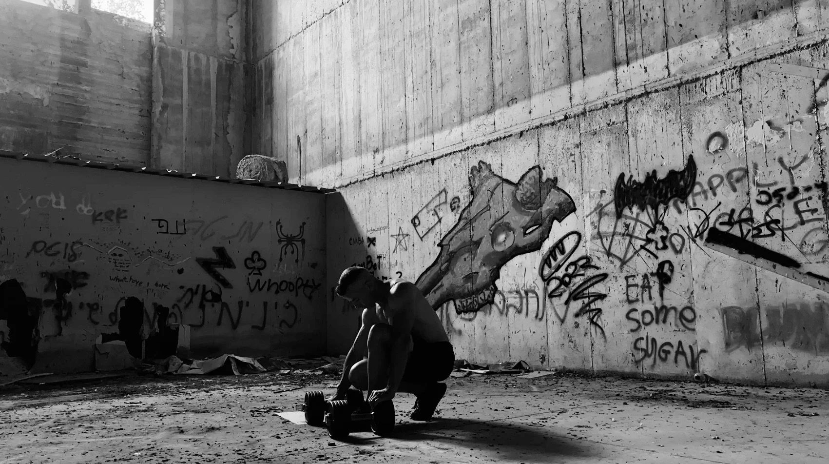 A shirtless man exercising with a dumbbell in a graffiti-covered, unfinished concrete room with debris on the floor.