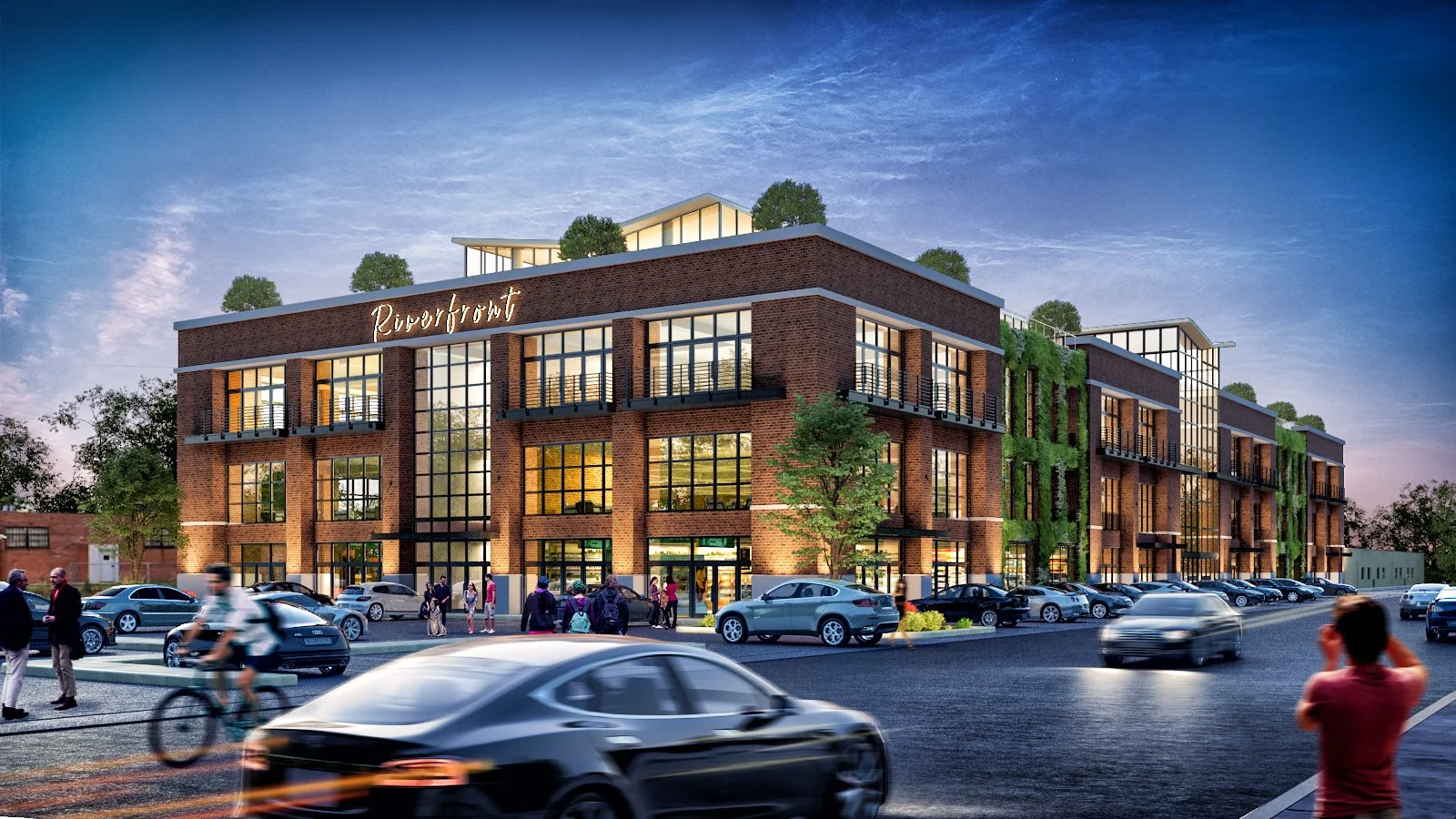 A modern brick building with large glass windows and the name 'Riverfront' illuminated on the facade. There are cars parked and driving in front of the building, and people walking on the sidewalk during dusk.