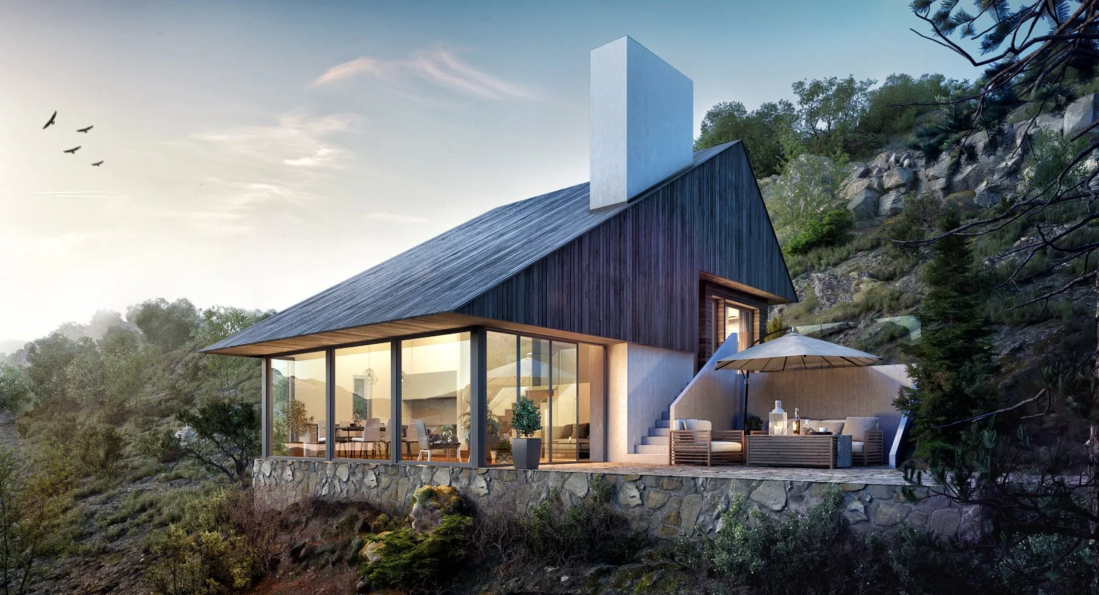 A modern house built on a hillside with large glass windows, a stone foundation, and a balcony with outdoor seating, surrounded by trees and rocks during sunset.