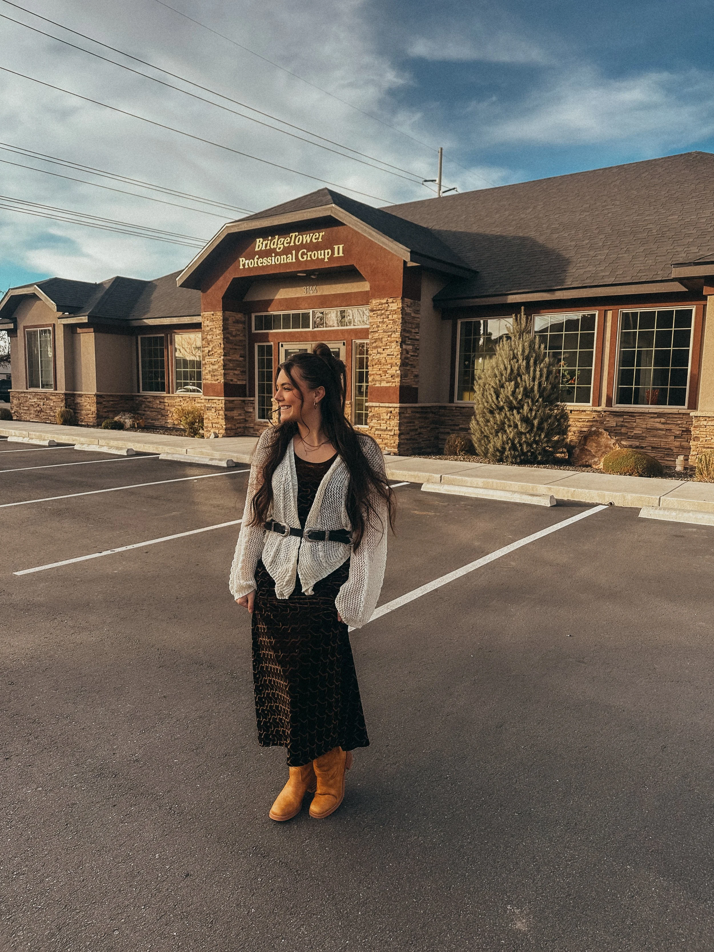A woman standing in a parking lot in front of a building with a sign that reads 'Bridge Tower Professional Group II.' The woman has long dark hair, is smiling, wearing a black dress, a white cardigan with a belt, and tan boots, and is looking to her left.