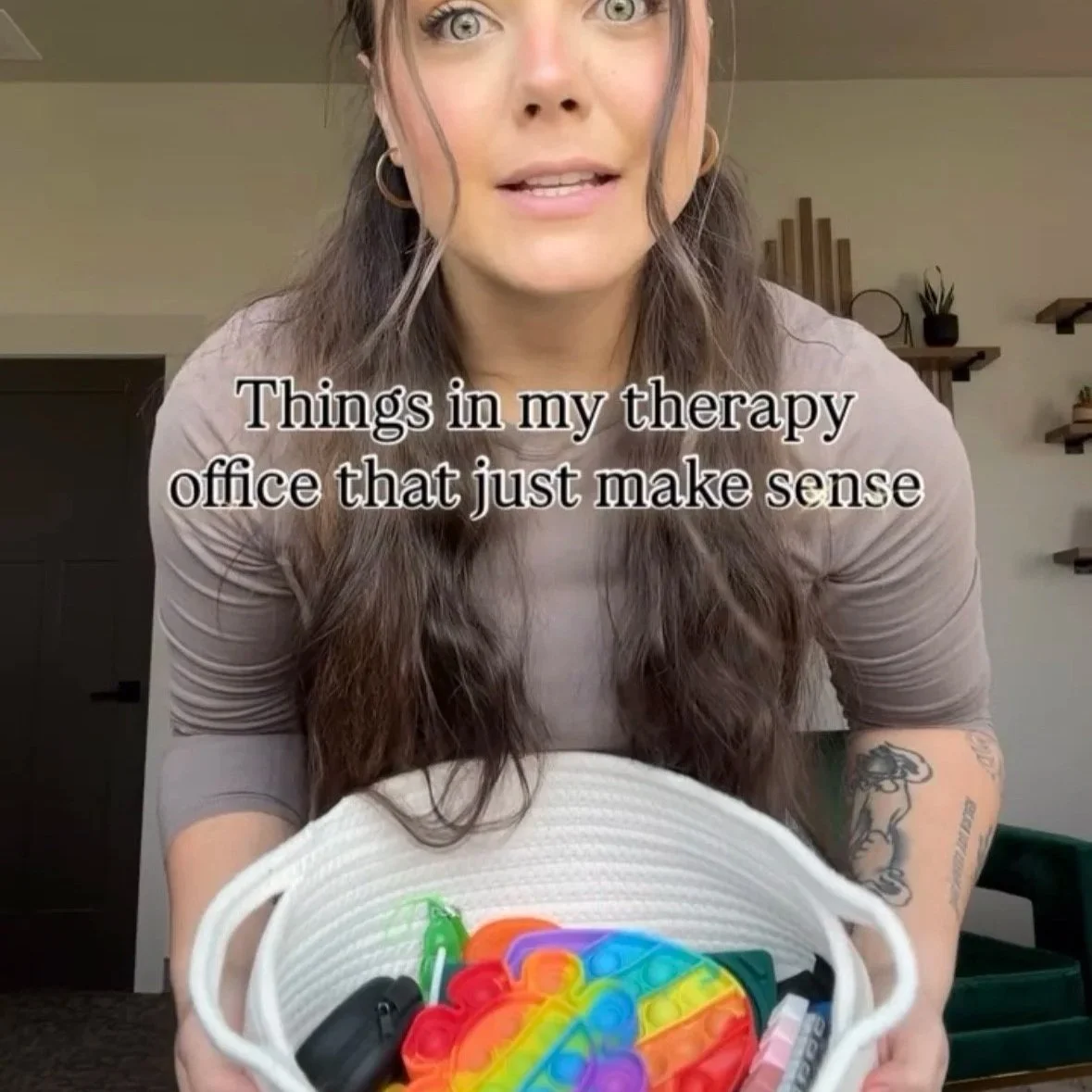 A woman with long brown hair holds a white basket filled with colorful sensory fidget toys, with a caption overlayed that reads 'Things in my therapy office that just make sense'.