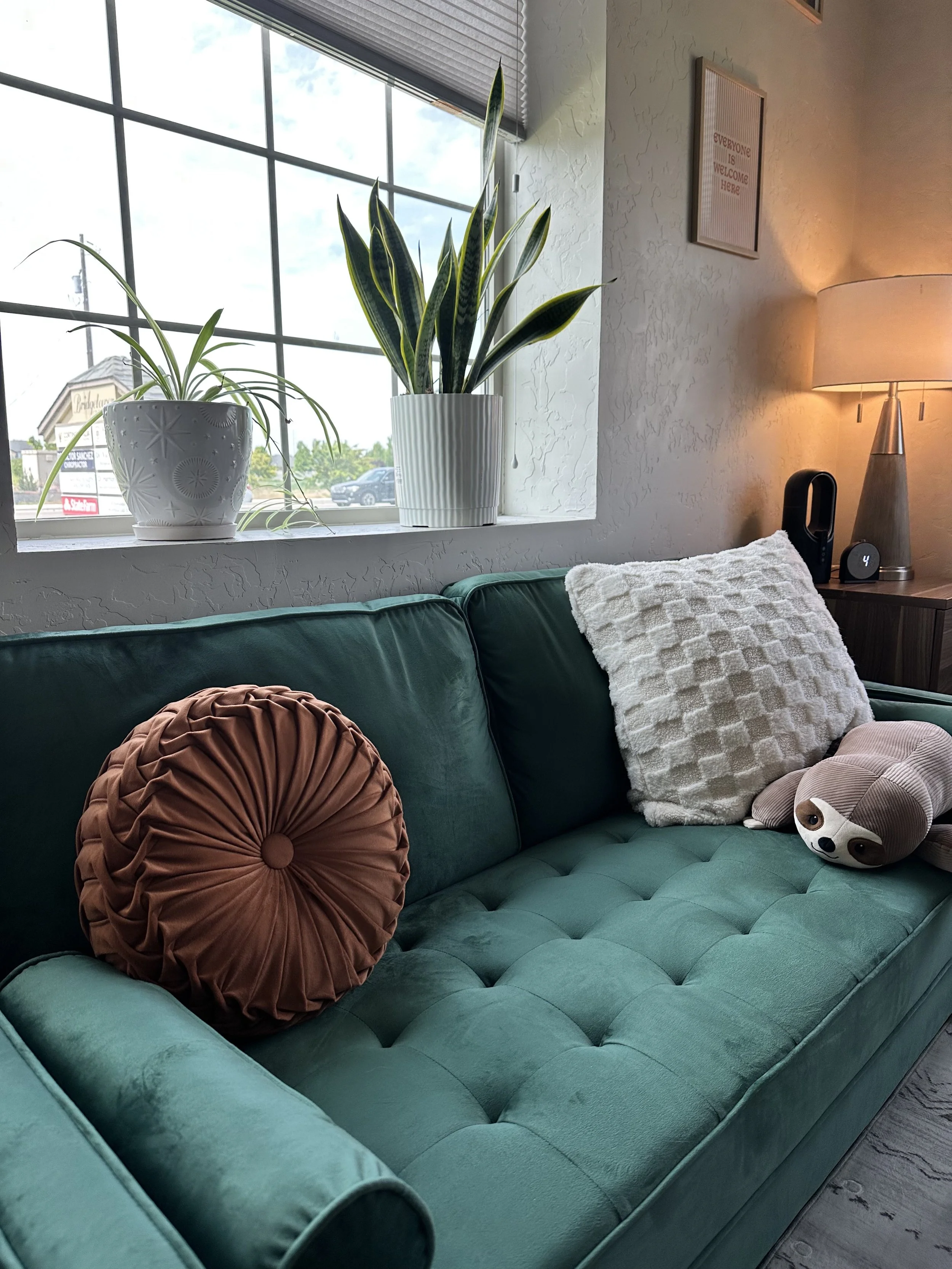 A teal tufted sofa with three pillows, a round velvet pillow, a fluffy white pillow, and a dog-shaped pillow, near a window with two potted plants and a wall lamp.