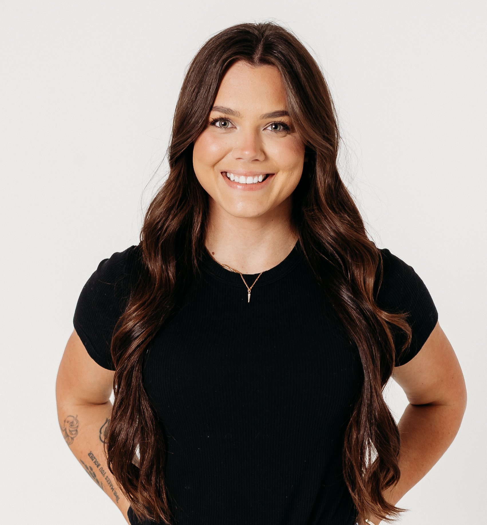 Close-up of a smiling woman with long, wavy, dark brown hair against a plain, light background.