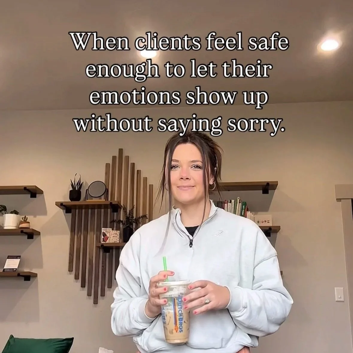 A woman smiling while holding a Starbucks iced coffee with a straw in a cozy living room with shelves and plants in the background. Text overlay: 'When clients feel safe enough to let their emotions show up without saying sorry.'