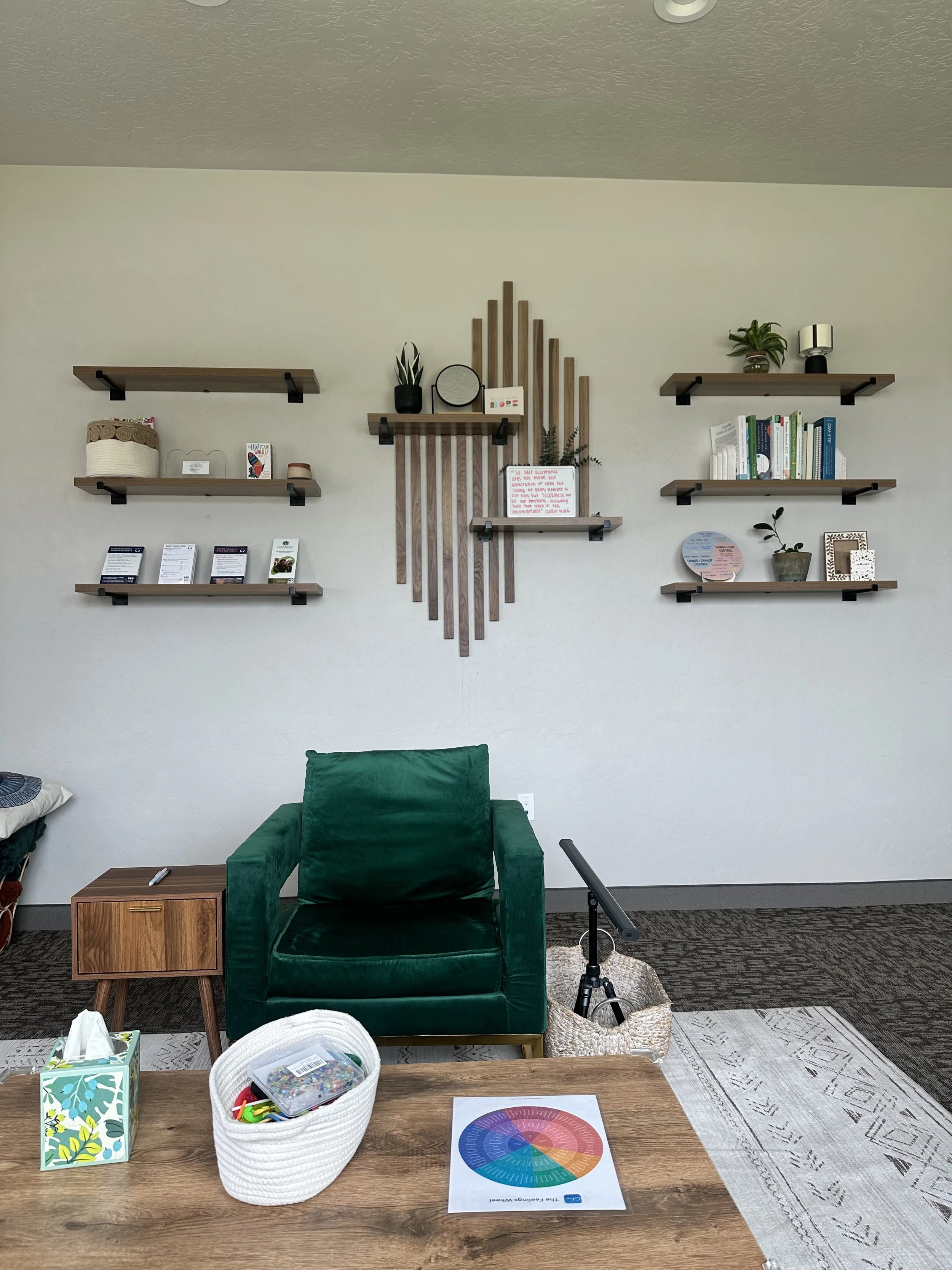 A room with a wooden table holding a color wheel chart, a tissue box, a white basket with supplies, and a pen. There is a green armchair, a small wooden side table, and a gray rug on a darker patterned carpet. The wall has six floating shelves with plants, books, and decorative items.