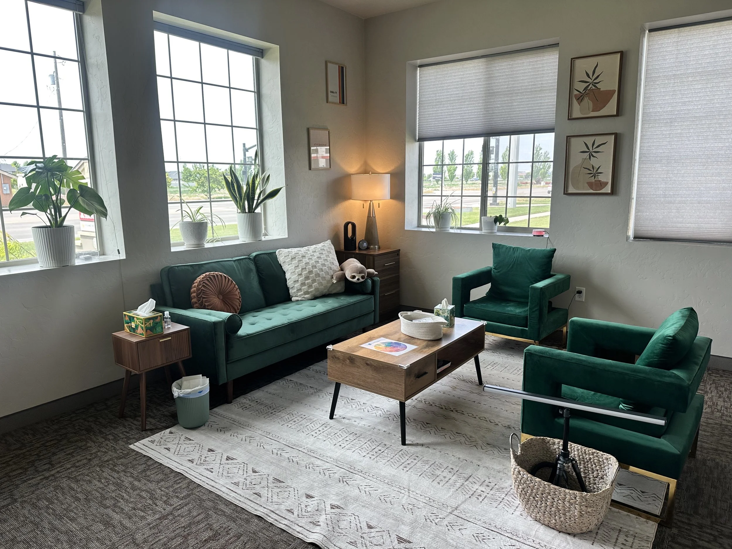 A cozy living room with large windows, green velvet sofas, a wooden coffee table, and various potted plants, with artwork on the walls and a small side table with a lamp.