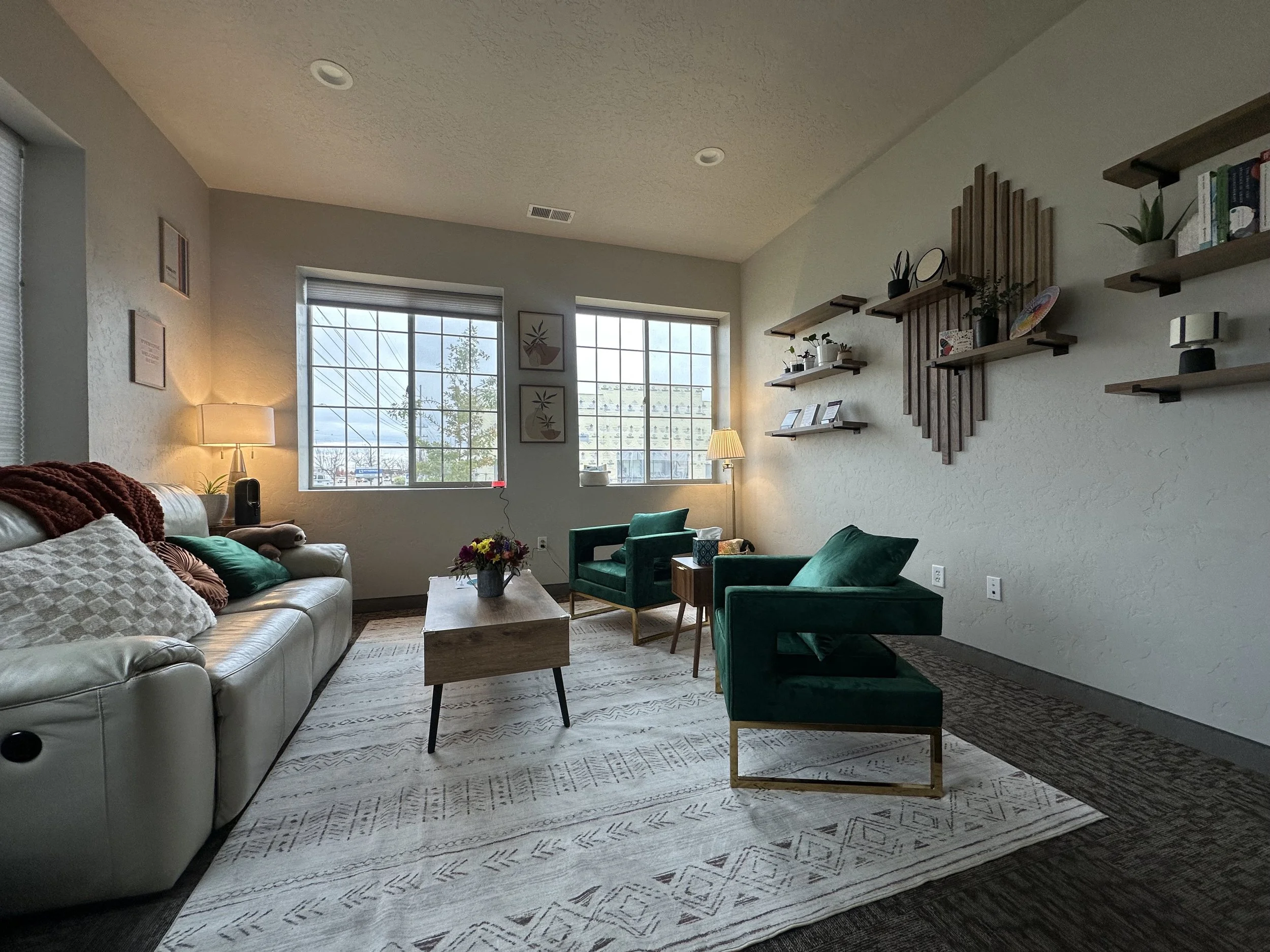 Living room with white sofa, green armchairs, wooden coffee table, bookshelves on the wall, and large windows with a view outside.
