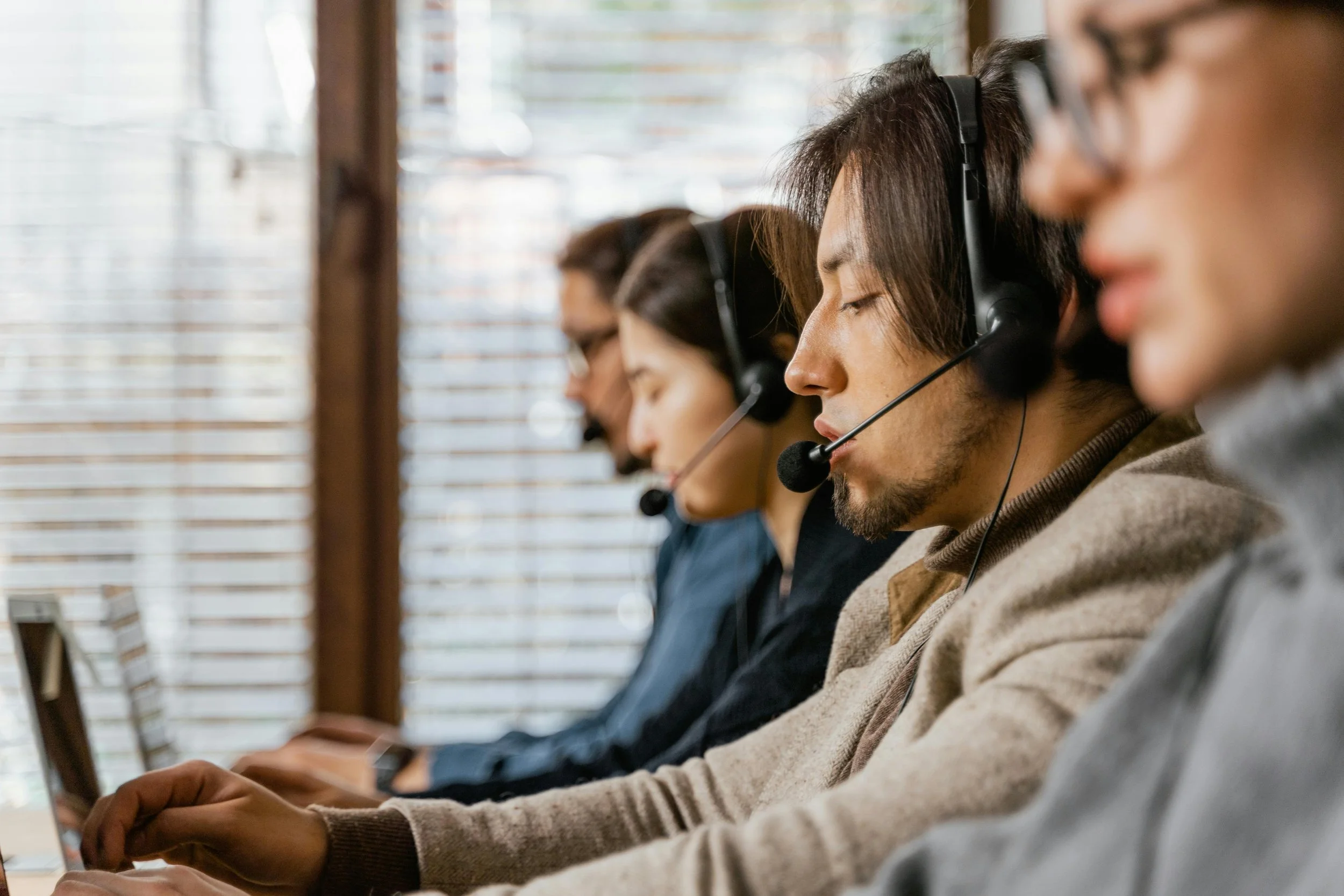 Four customer service representatives wearing headsets with microphones, sitting at desks with computers, working in an office environment.