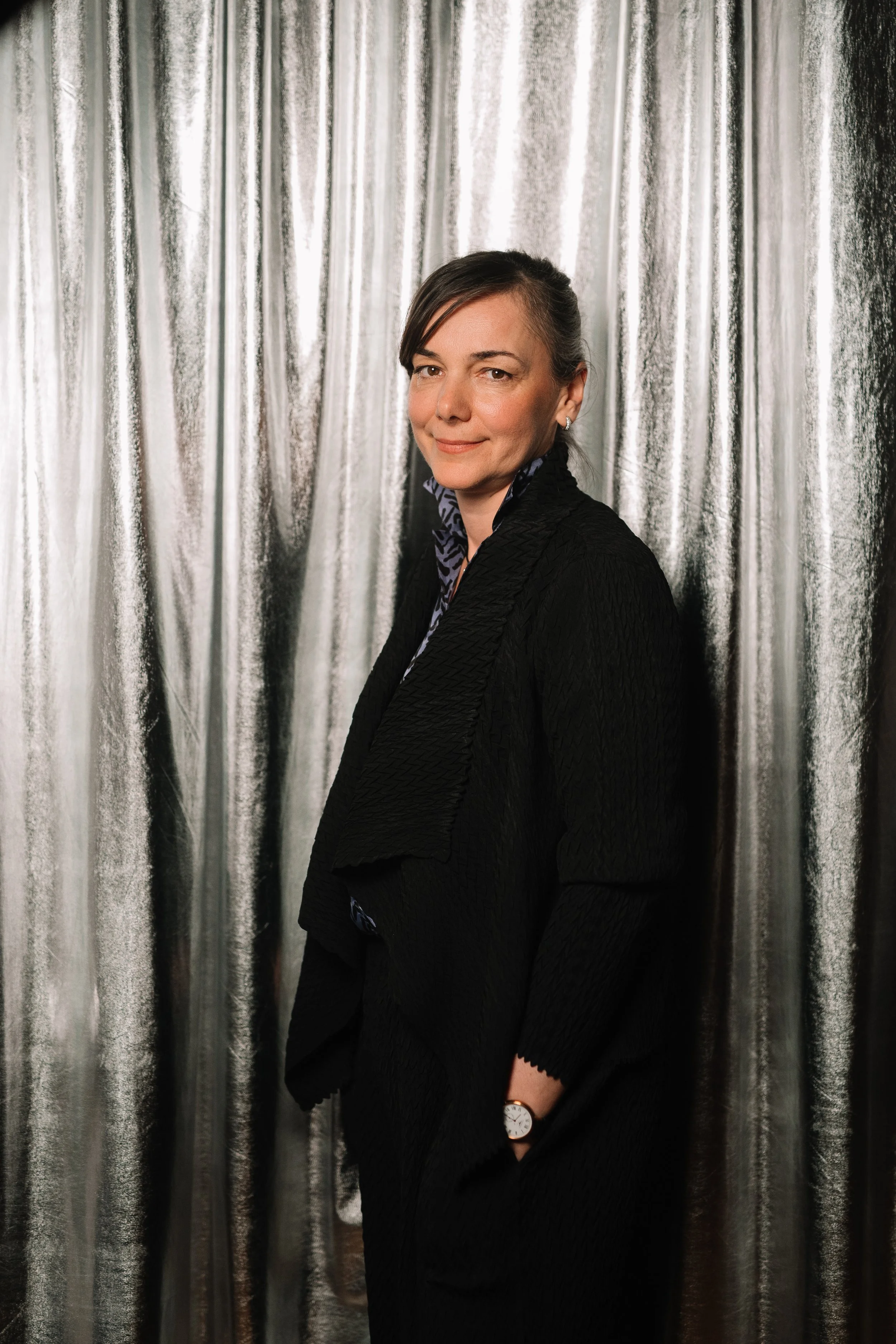 A woman in a black textured jacket and patterned blouse standing in front of a metallic curtain.