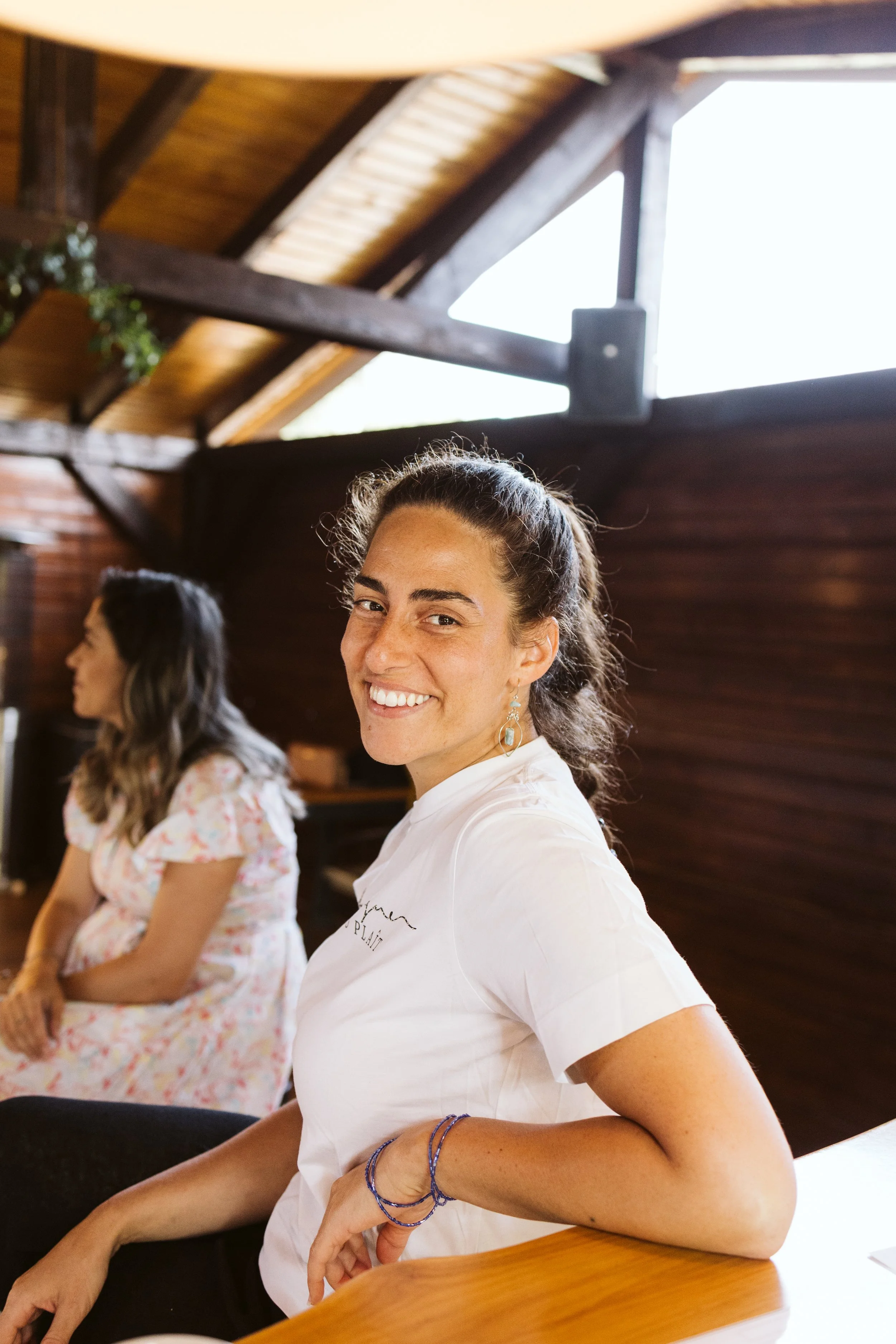 A woman smiling at the camera, sitting inside with another woman in the background. The setting appears to be a wooden cabin or barn with natural light coming through the windows.