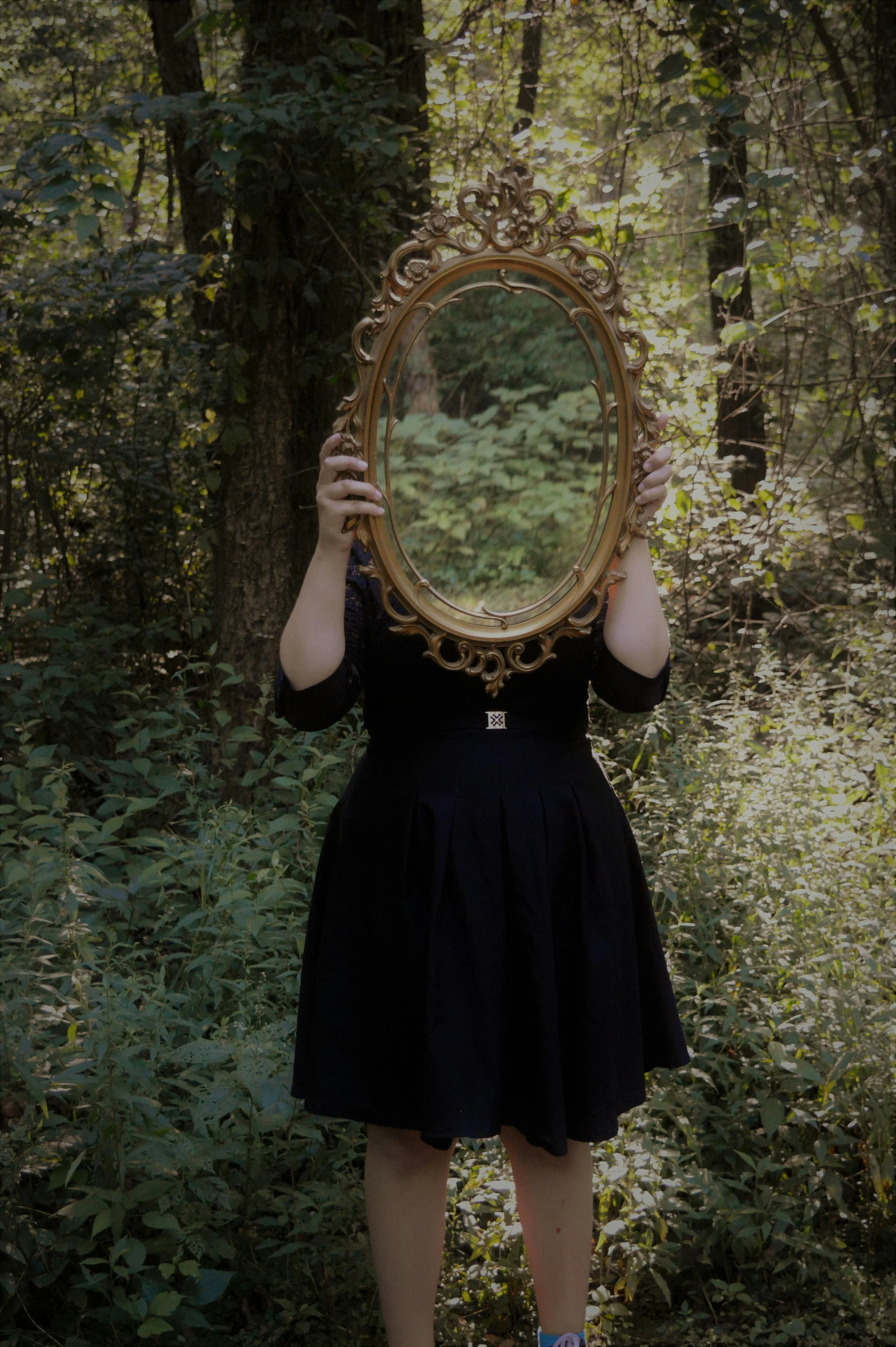 Person in black dress standing in a forest, holding an ornate oval mirror in front of their face, reflecting the forest behind.