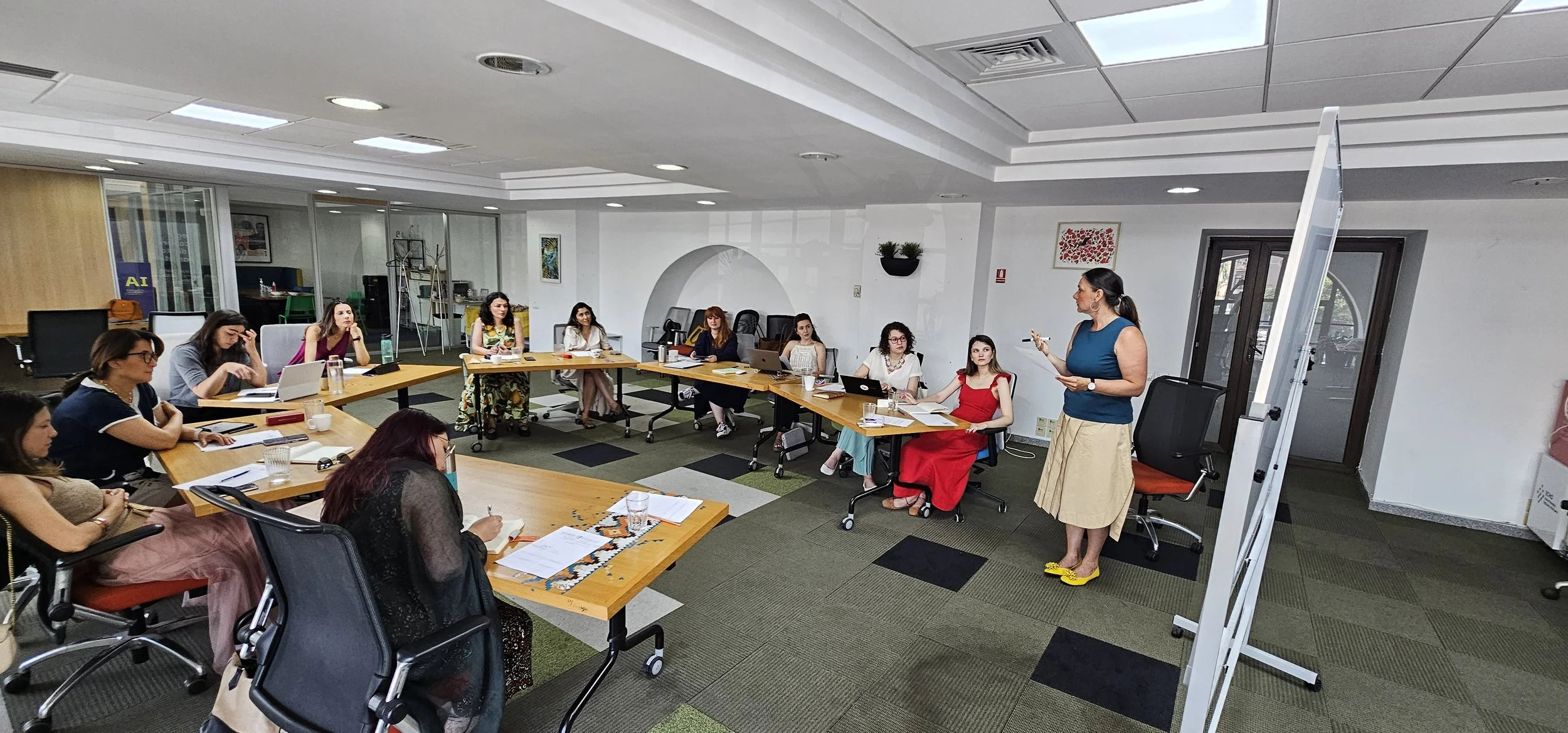 A group of women attending a meeting in a conference room, with a woman standing and speaking at the front, holding a notepad and wearing yellow shoes.