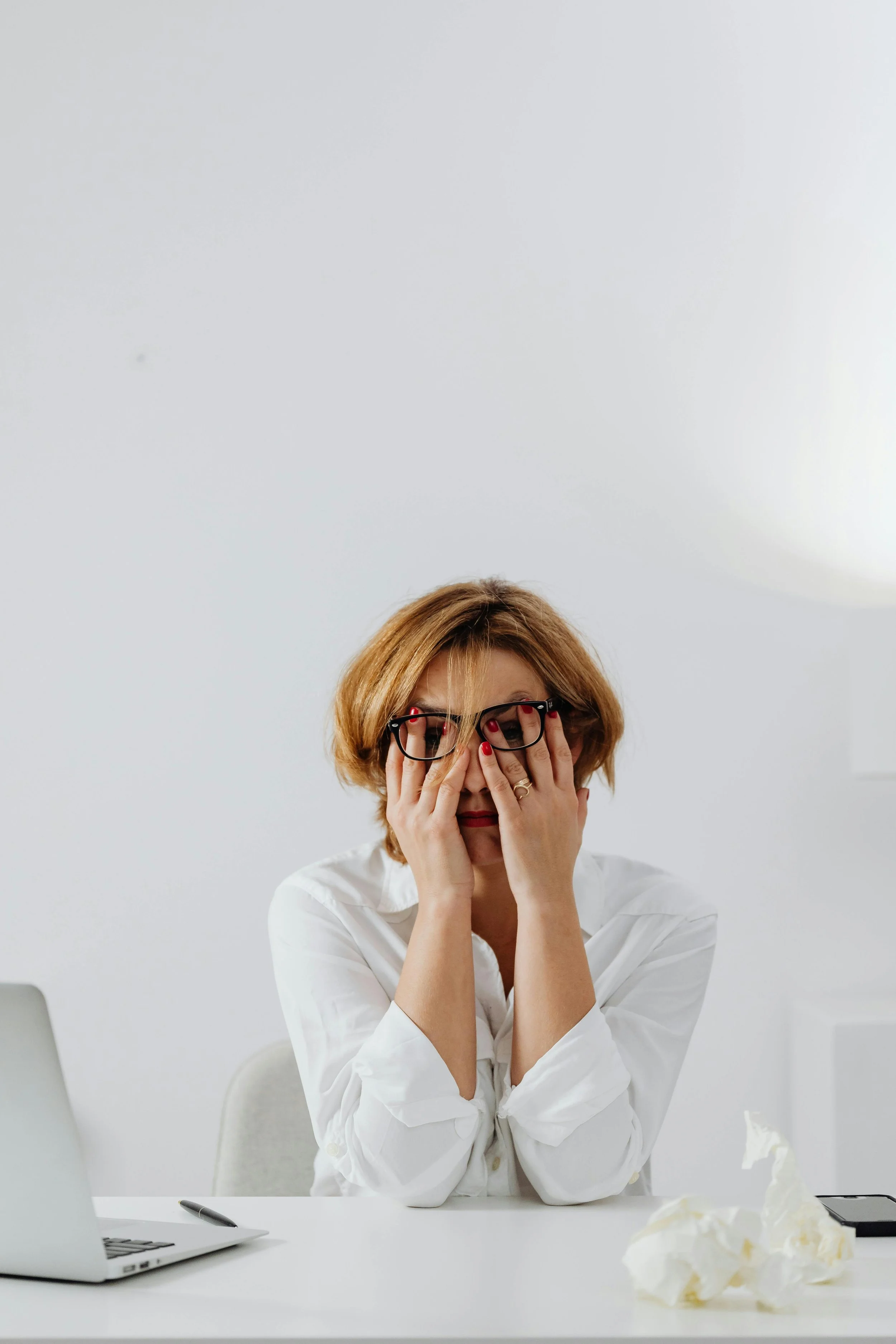 Woman with glasses covering her face with her hands sitting at a white desk, with a laptop, a pen, a crumpled tissue, and a phone, against a plain white background, appearing stressed or overwhelmed.