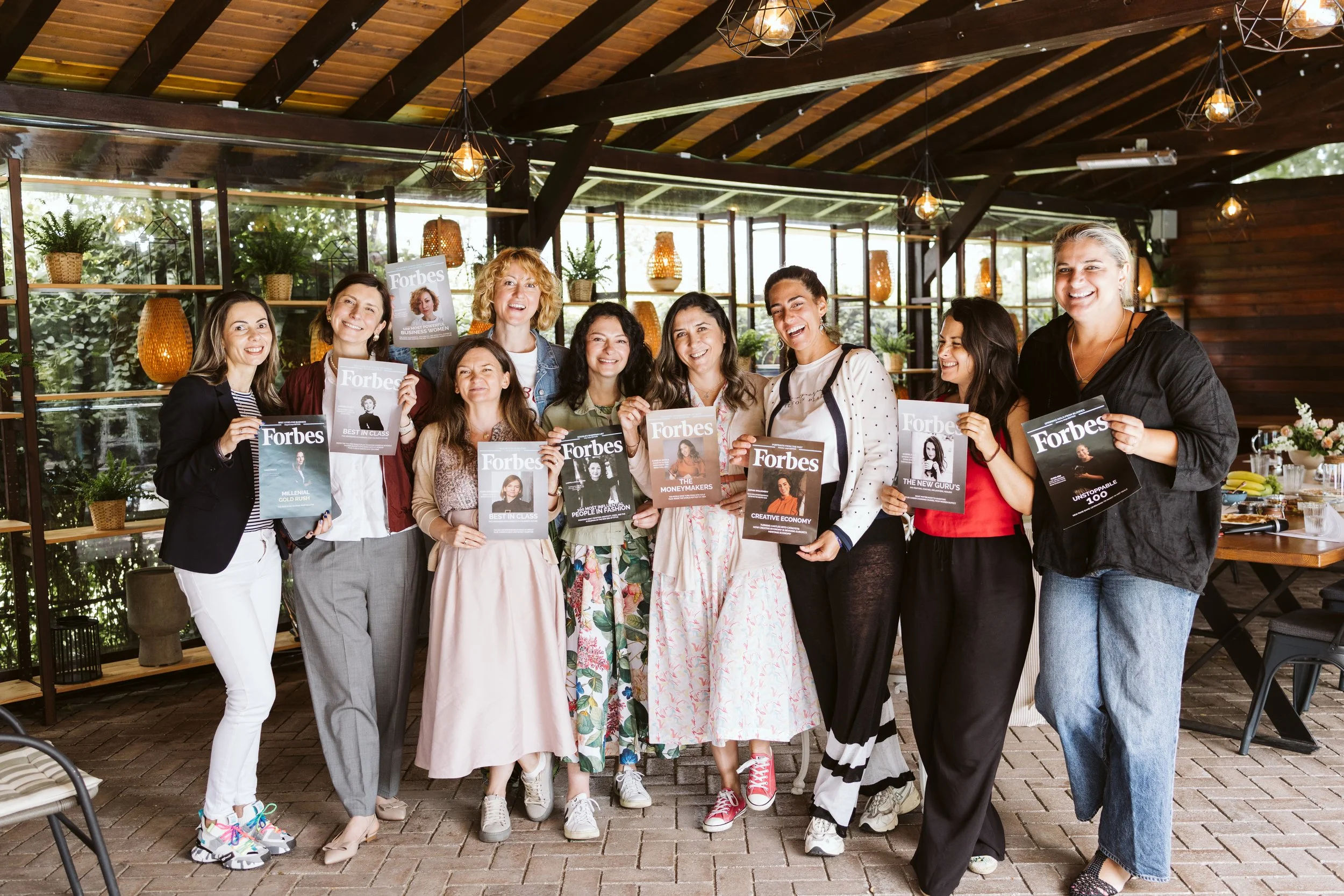 Group of women holding Forbes magazine covers in a bright, indoor space with wooden ceiling and large windows.