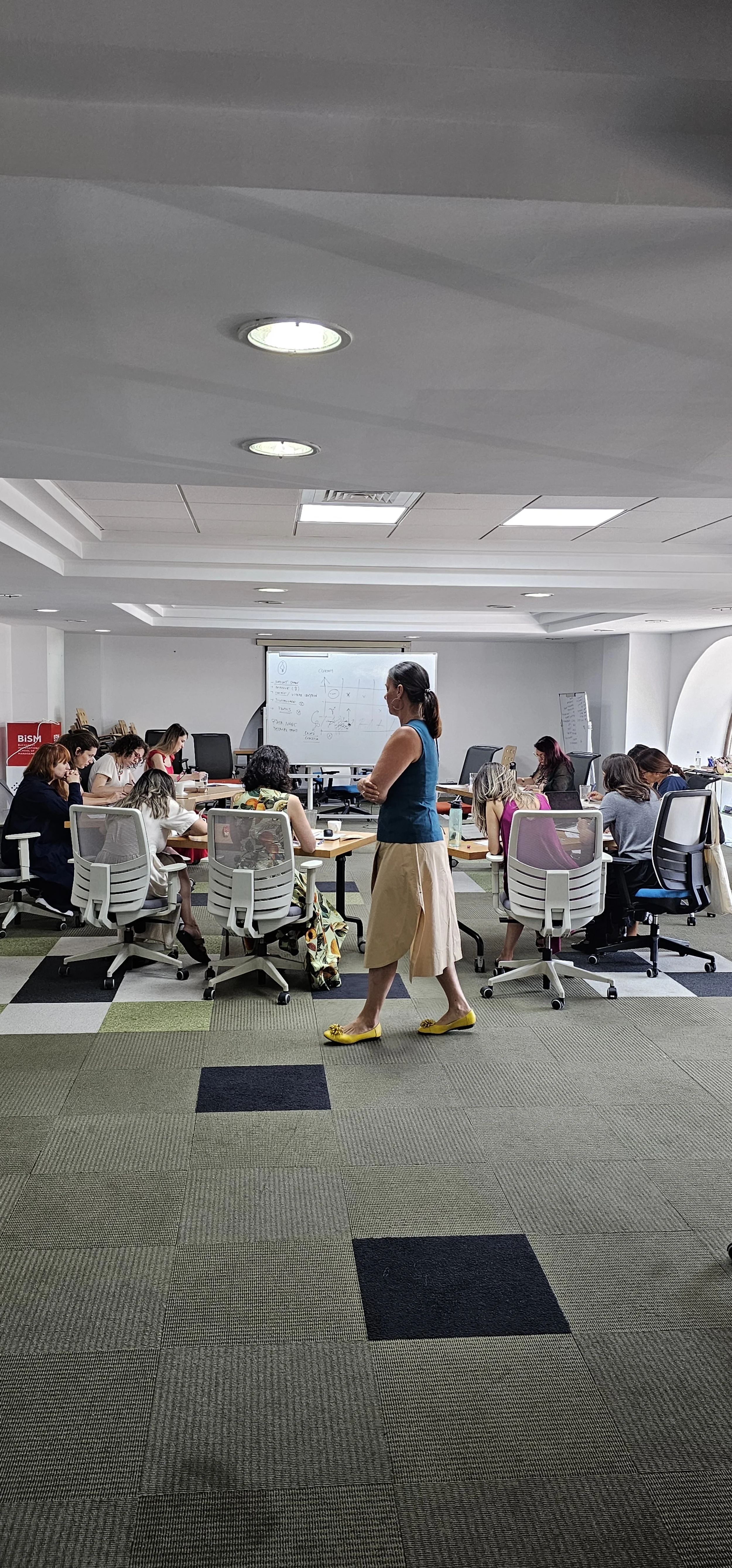 A classroom or conference room with a group of women seated around a large table, a woman in a blue top and beige skirt standing near the middle, facing the women, with a whiteboard and a monitor at the front of the room, and geometric diagrams on the whiteboard.