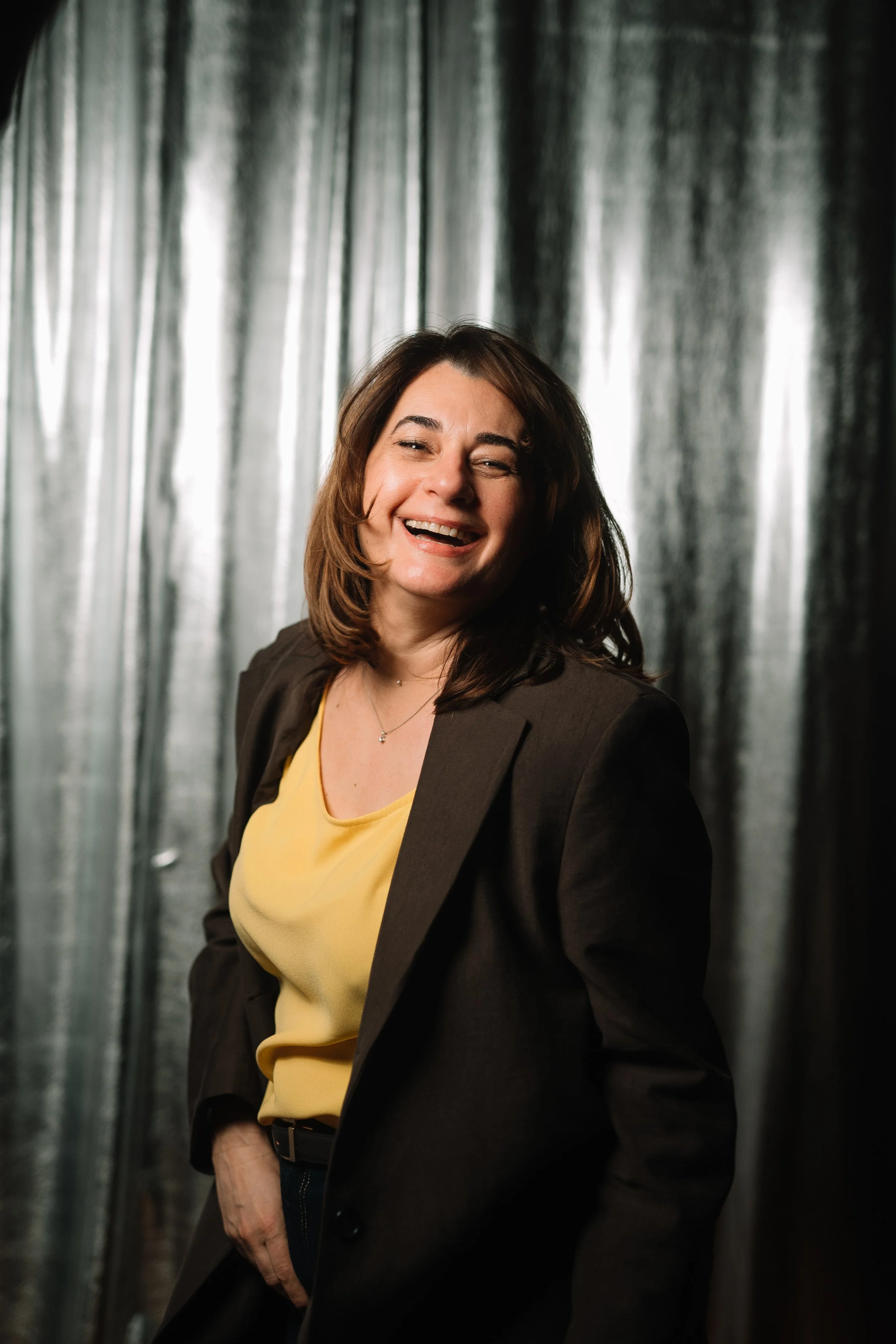 A woman with shoulder-length brown hair smiling and winking, wearing a yellow top and a dark blazer, standing in front of metallic curtains.